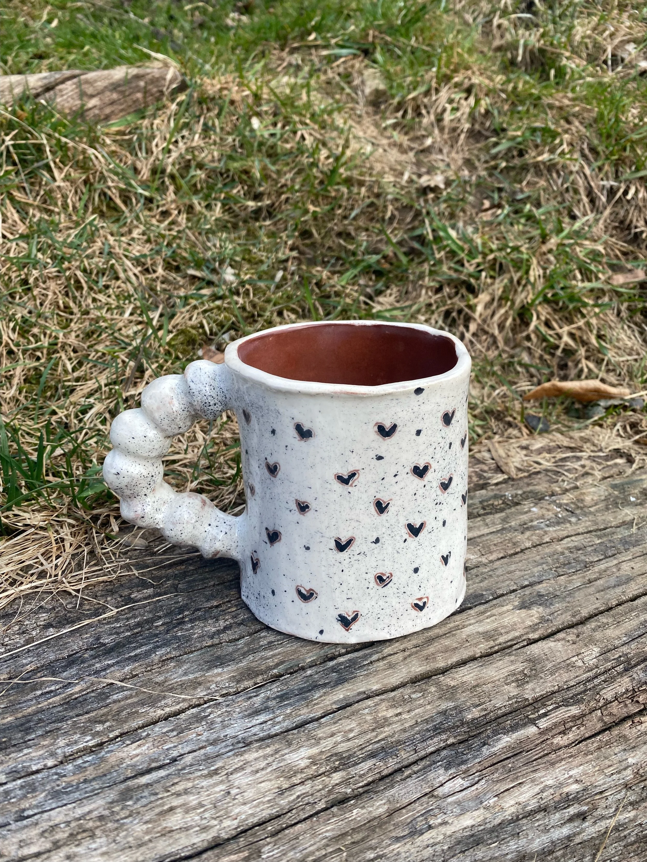 Tasse en céramique blanche à petits cœurs noirs, posée sur une vieille planche en bois, avec de l'herbe et des plantes en arrière-plan.