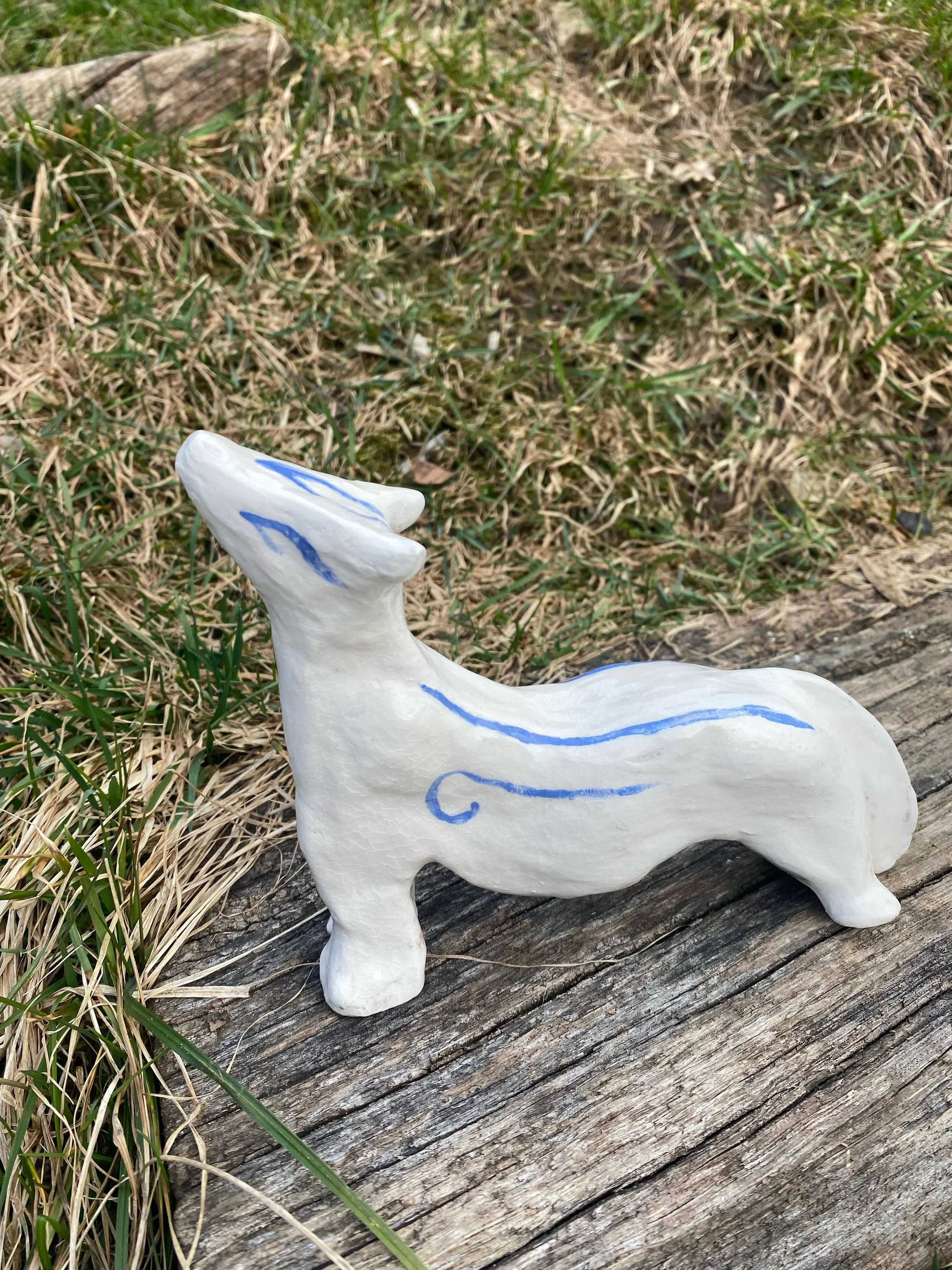 Un sculpture en céramique blanche représentant un animal fantastique ou une créature mythique, avec des lignes bleues décoratives sur le corps et la tête, placée sur un vieux bois à l'extérieur, entourée d'herbe.