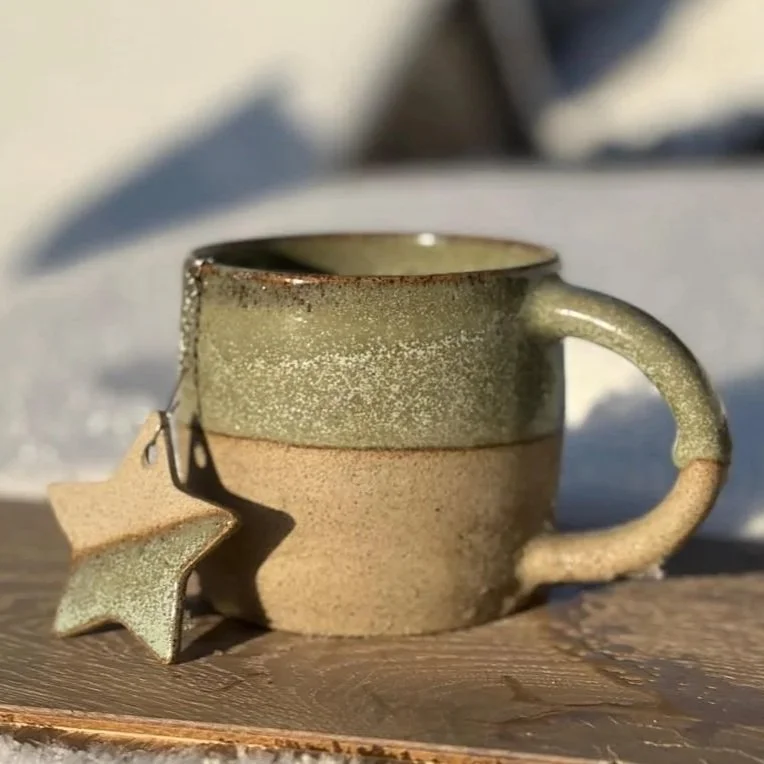 Une tasse en grès verte et beige avec une boule à thé accroché par une étoile en grès, posée sur une surface en bois, avec de la neige en arrière-plan.
