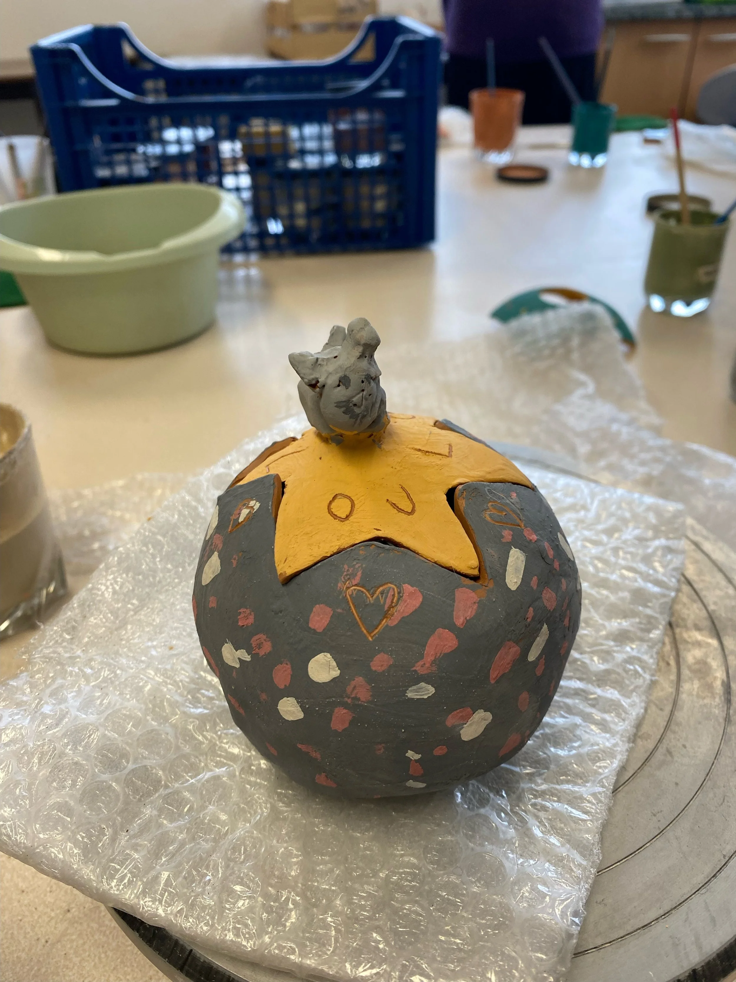 Une décoration en forme de boule peinte avec des motifs en cœur, en taches rosées et blanches, avec une étoile jaune sur le dessus, et une petite sculpture en forme de chat ou chaton en haut, posée sur une surface recouverte de bulle plastique, dans un atelier.