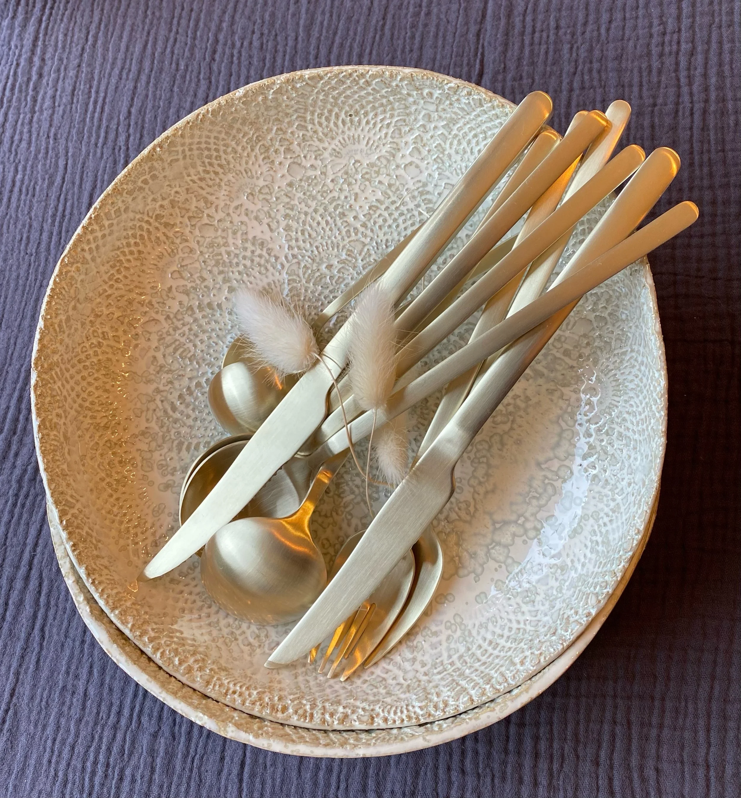 Assiette blanche avec plusieurs cuillères, fourchettes et couteaux en métal, décorée avec de petites plumes blanches, placé sur un tissu violet.