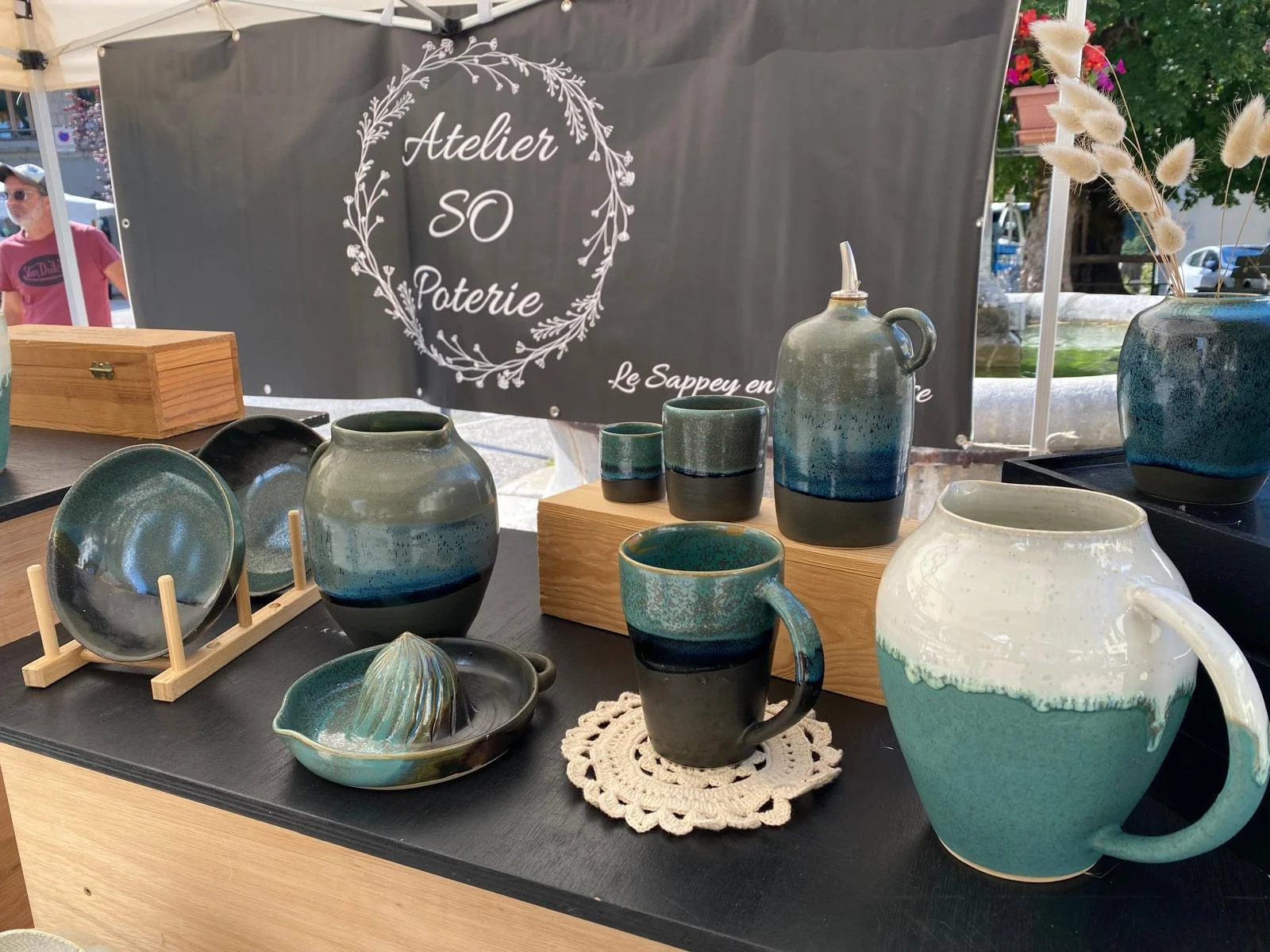 Vase, tasses, et plat en céramique artisanale de couleur verte et bleue sur une table, avec un panneau publicitaire en arrière-plan pour un atelier de poterie.