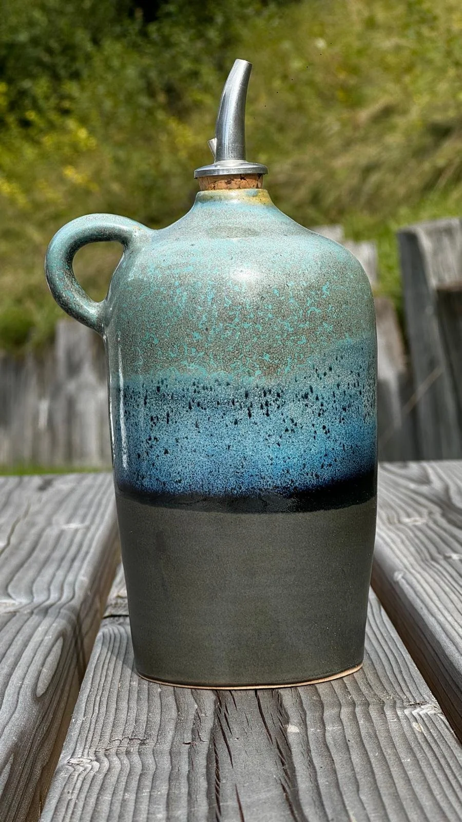 Un huillier en céramique avec un couvercle métallique, posée sur une table en bois, en plein air avec un fond de feuillage vert. L'huillier est colorée en dégradé de bleu, vert et noir.