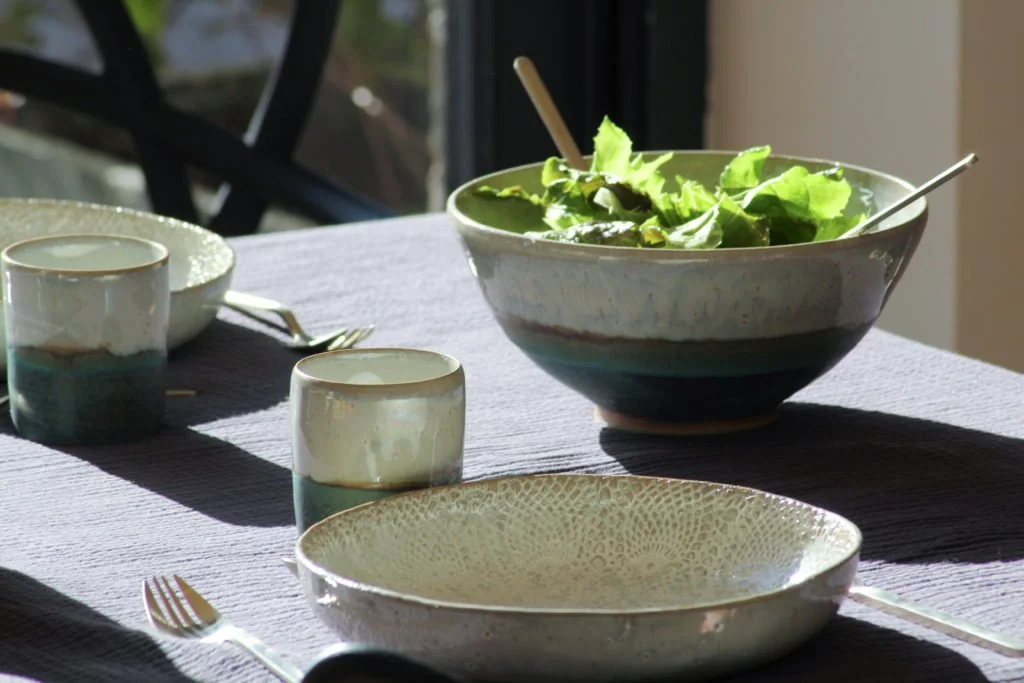 Une table de repas avec un saladier rempli de feuilles de salade, deux petits bols, deux fourchettes, une grande assiette creuse et deux verres. La scène est éclairée par la lumière naturelle.