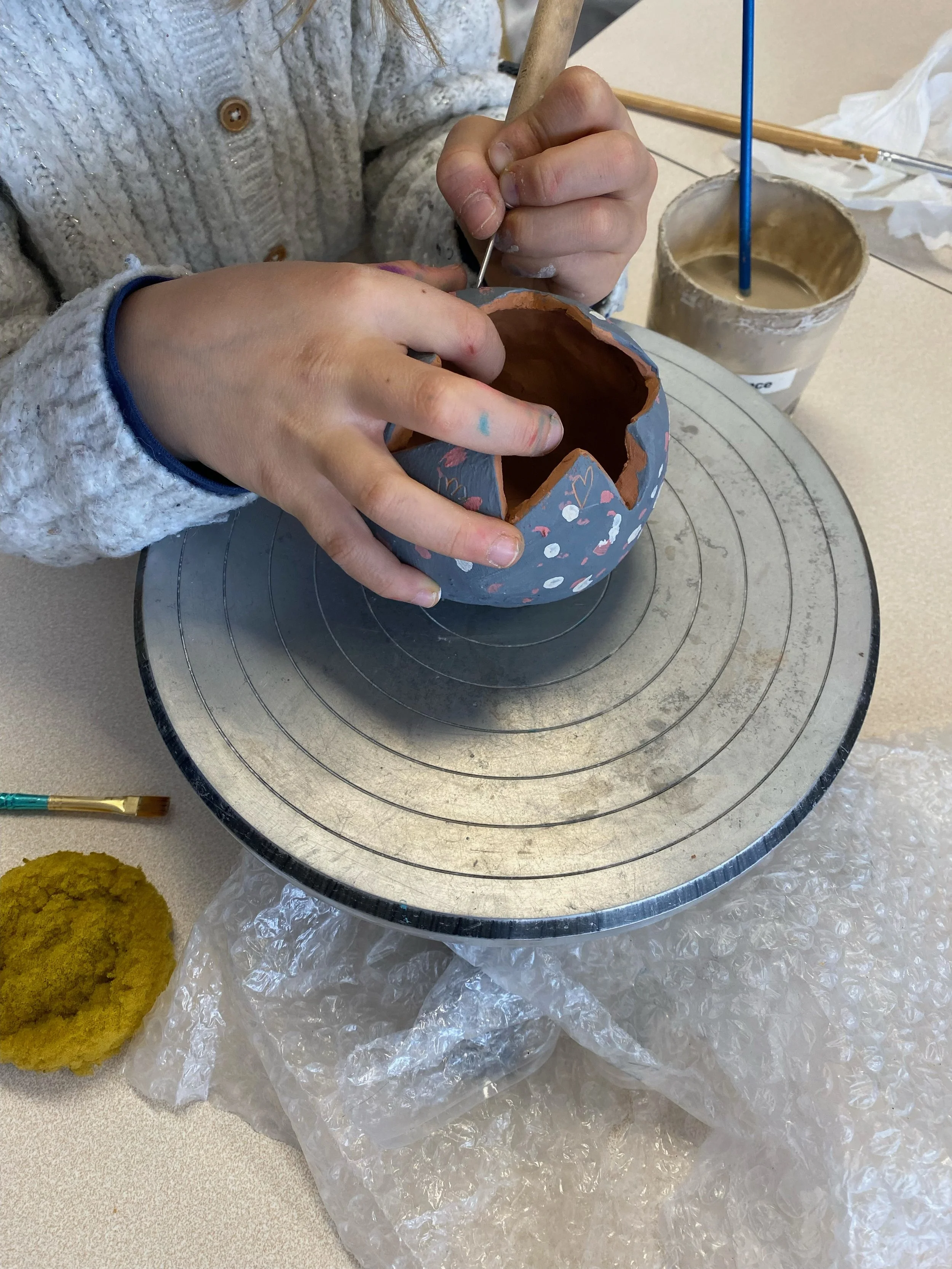 Une personne en train de peindre une boule en céramique de couleur grise avec des motifs colorés. La personne utilise un petit pinceau pour la décoration et travaille sur une surface tournante. Il y a un pot de peinture et une éponge à proximité.