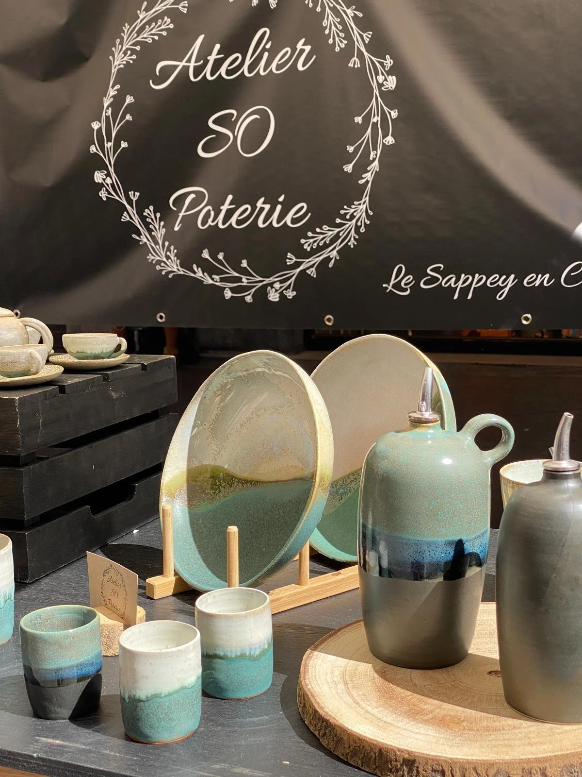 Vase, tasses, assiettes et huilliers en céramique colorée, exposés dans un marché de poterie. Un panneau noir avec inscription en français "Atelier SO Poterie".