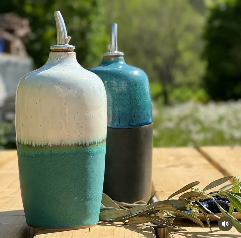 Deux huilliers en céramique avec bec verseur en métal, placées sur une table en bois, avec des branches d'olivier à côté, en plein air avec un fond d'arbres verts.