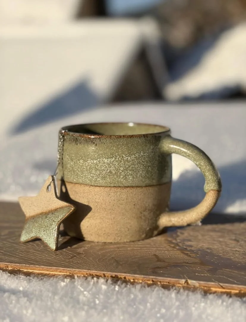 Une tasse en céramique verte et beige avec une étoile en céramique suspendue à la anse permettant de tenir une boule à thé à l'intérieur de la tasse, posée sur une surface en bois dans la neige, avec un fond flou.