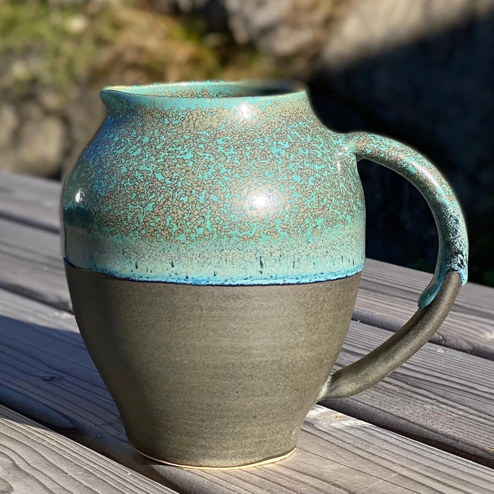 Une cruche en céramique avec une moitié inférieure en couleur gris foncé et une moitié supérieure décorée de motifs bleus et dorés, placée sur une surface en bois.