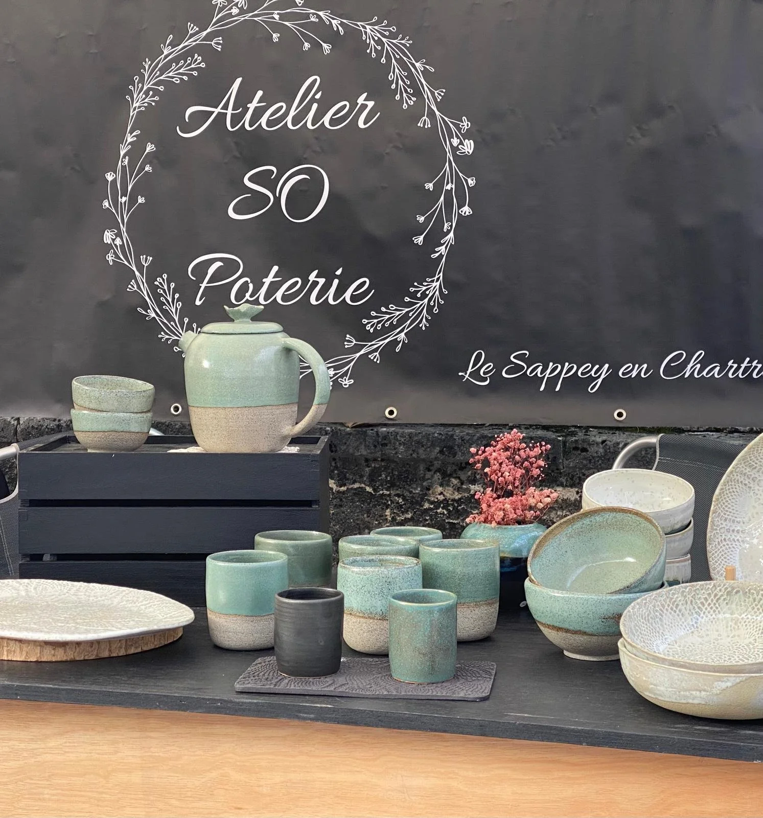 Vitrine avec des pièces en céramique, y compris des bols, tasses et un mug, devant un panneau indiquant 'Atelier SO Poterie' à Le Sappey en Chartreuse.