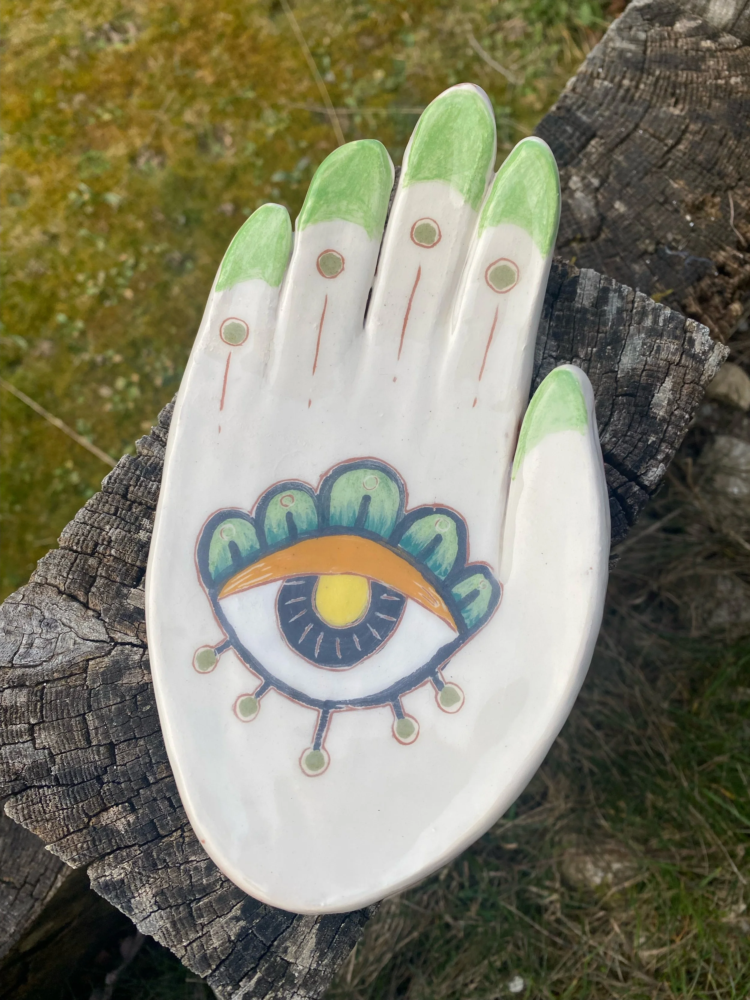 Une plaque décorative en forme de main avec un motif d'œil stylisé au centre, ornée de couleurs vives telles que le vert, le jaune et l'orange, posée sur une surface en bois.