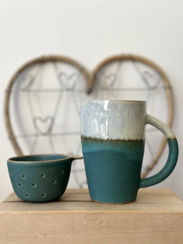 Une tasse en céramique bleue et blanche et un petit bol en céramique bleue sur une surface en bois, avec un décor en forme de cœur en fil métallique en arrière-plan.