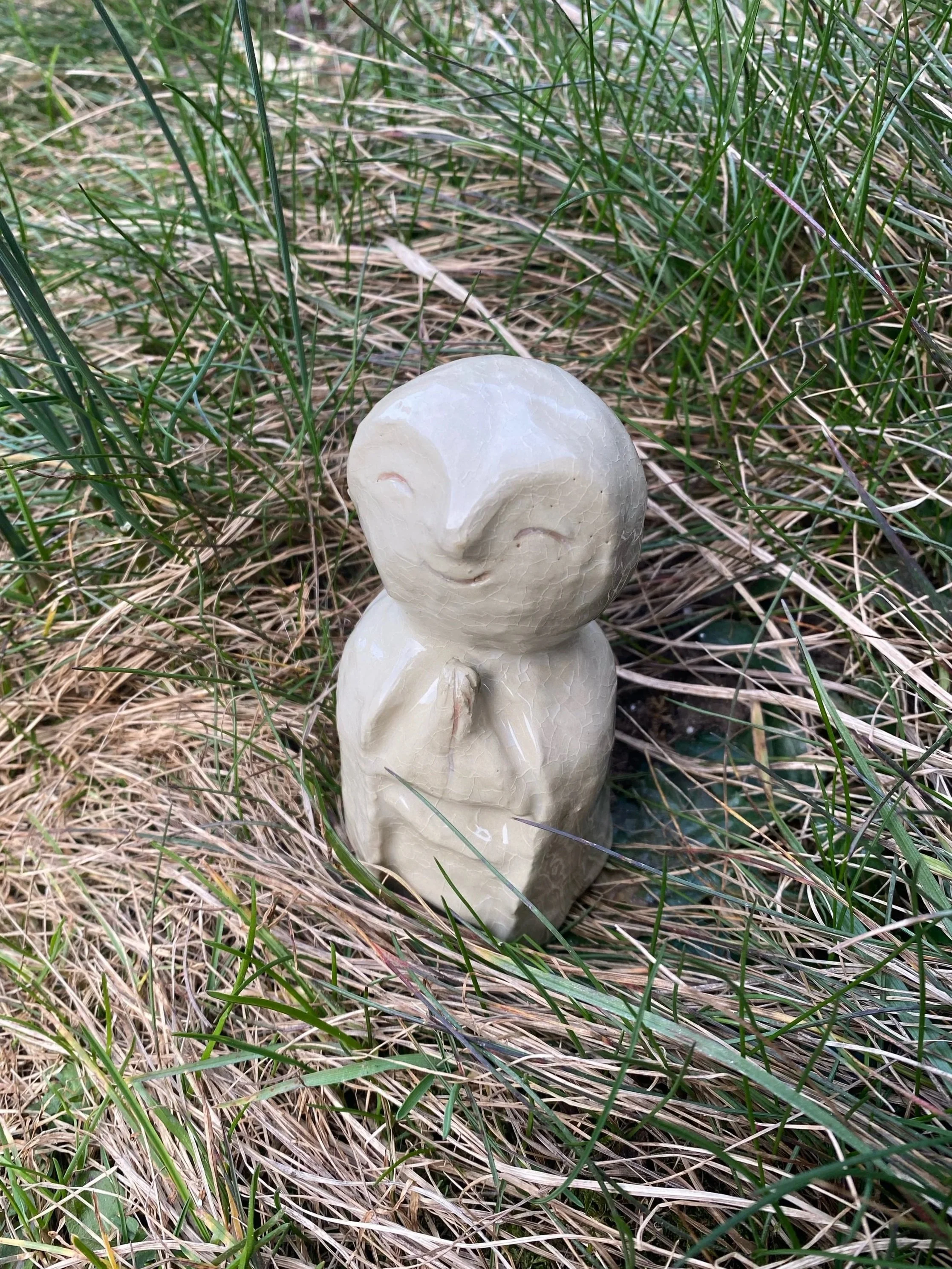 Statue en forme de personnage avec les mains jointes, souriant, entourée d'herbe et de plantes