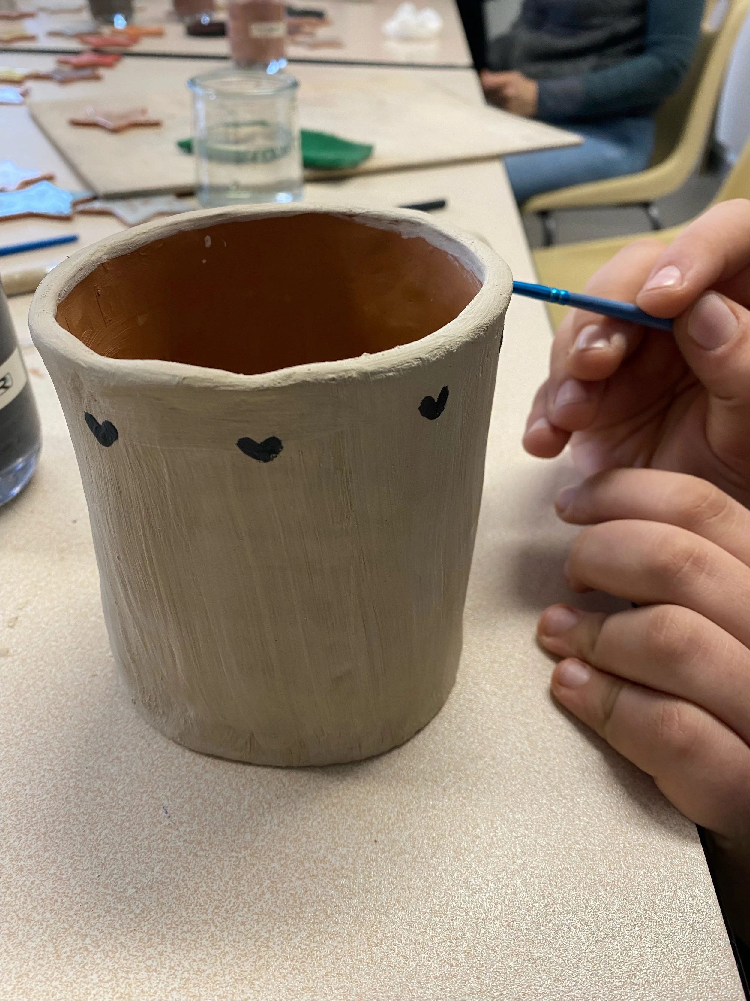 Une personne peignant une poterie en terre cuite beige avec de petits cœurs noirs autour de la surface, assise à une table.