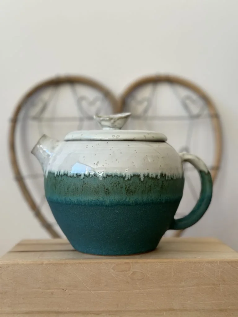 Une théière en céramique verte et blanche posée sur un support en bois, avec un meuble en bois clair en arrière-plan.
