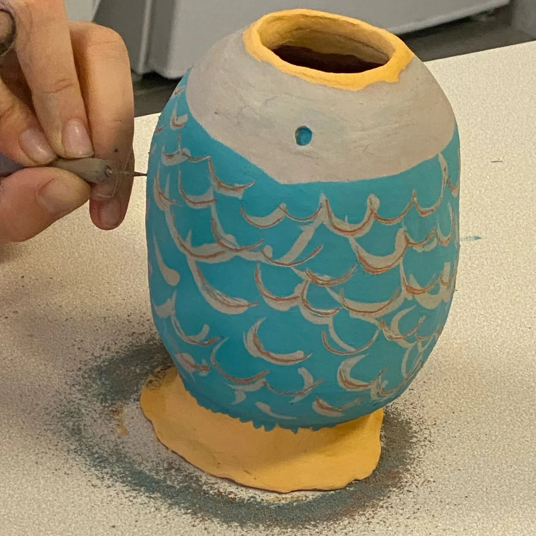 Une personne peignant un vase en terre cuite en forme de poisson avec des motifs d'écailles en utilisant de la peinture bleue et blanche.