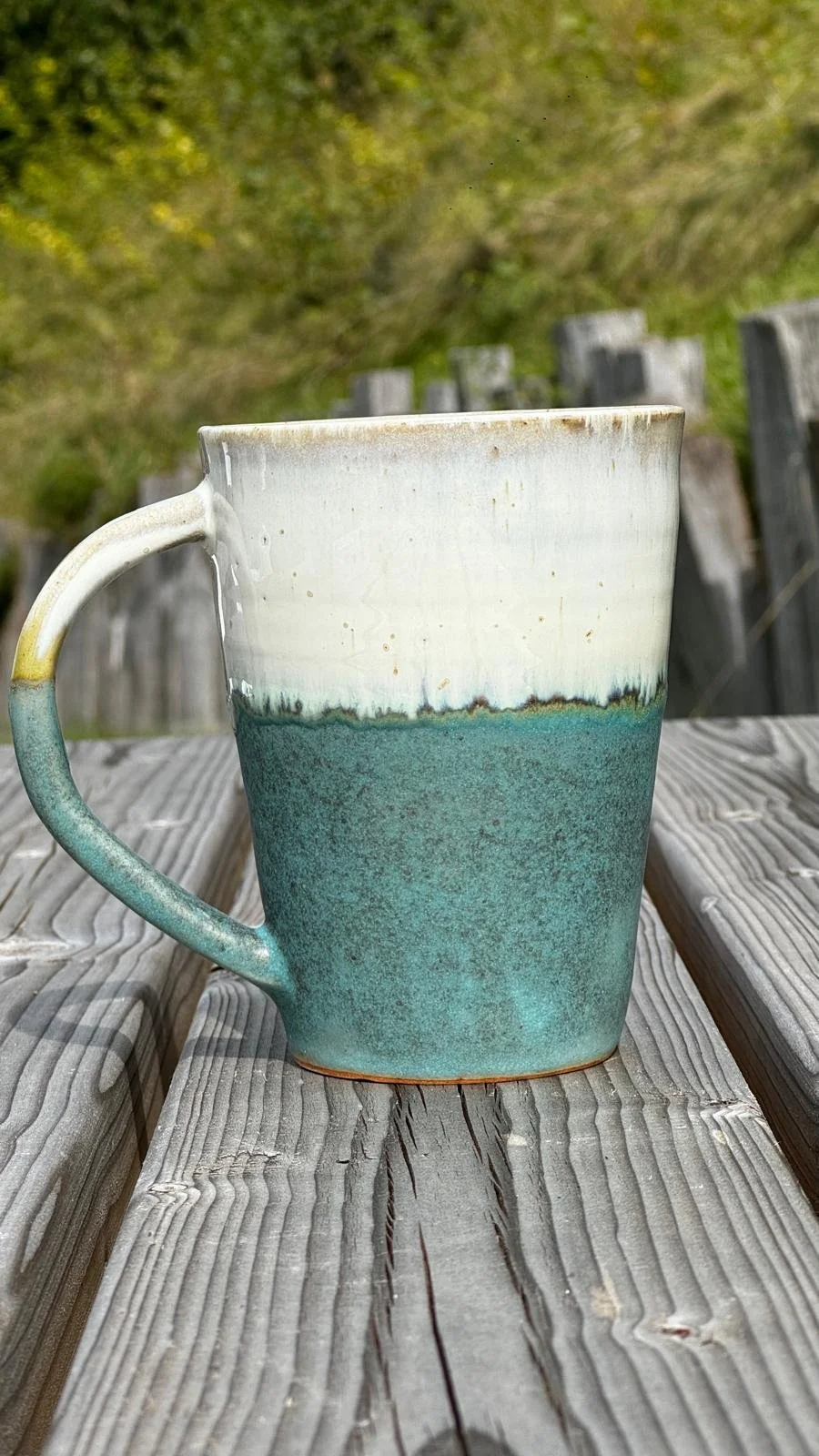 Une tasse en céramique verte et blanche posée sur une table en bois dehors, avec un fond flou d'arbres et de verdure.