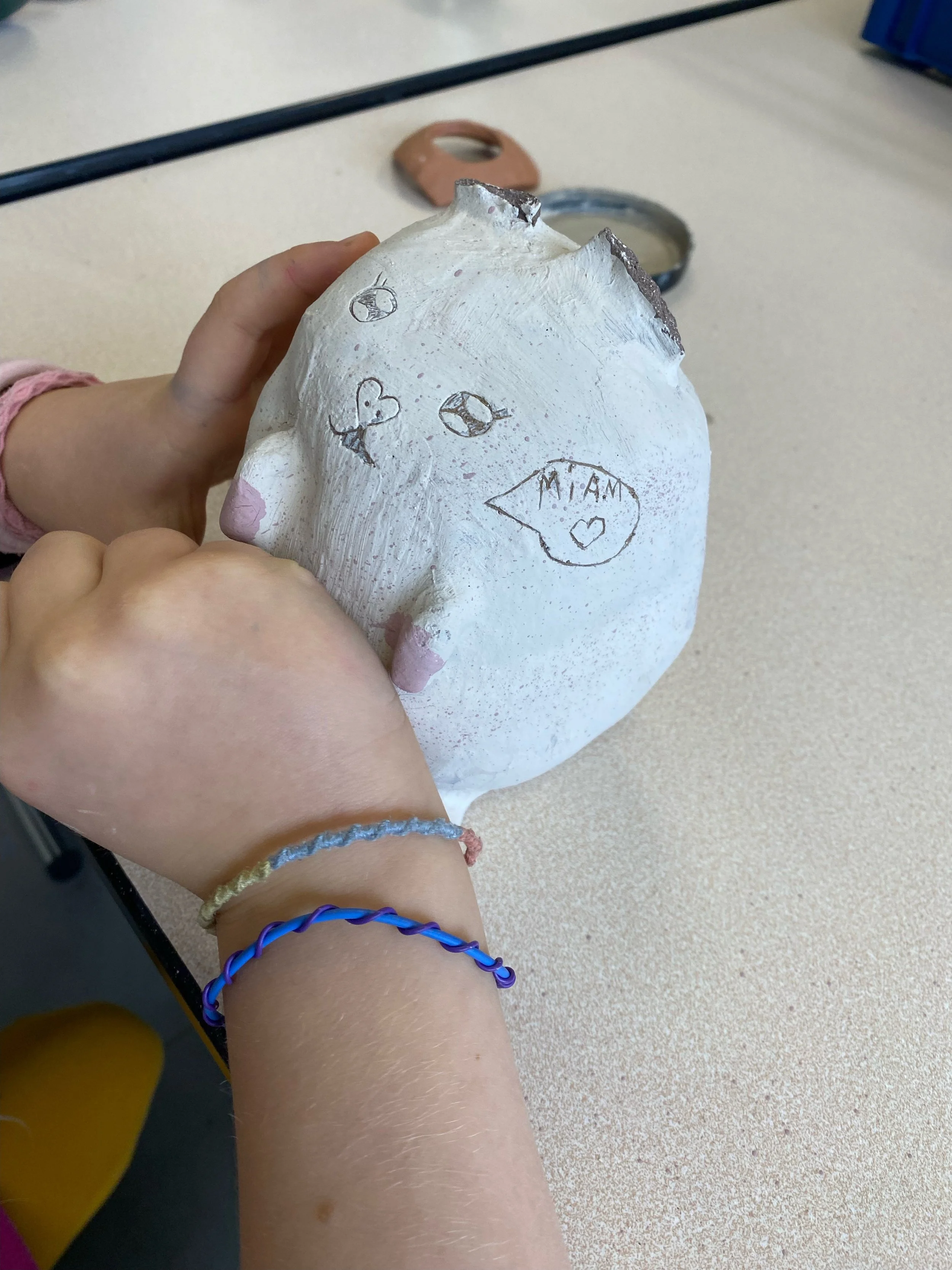 Enfants tenant une petite sculpture en céramique blanche avec des dessins de visages, un cœur, et une bulle de dialogue contenant 'MIAM' avec un cœur. La sculpture est dans un atelier.