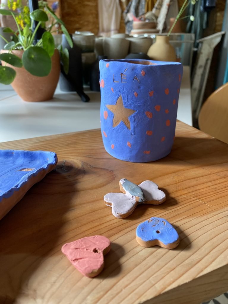 Un vase en argile peint en bleu avec des décorations en forme d'étoile et de points, entouré de petits biscuits décorés en forme de papillon et de nuage sur une table en bois, avec un fond de plantes et meubles