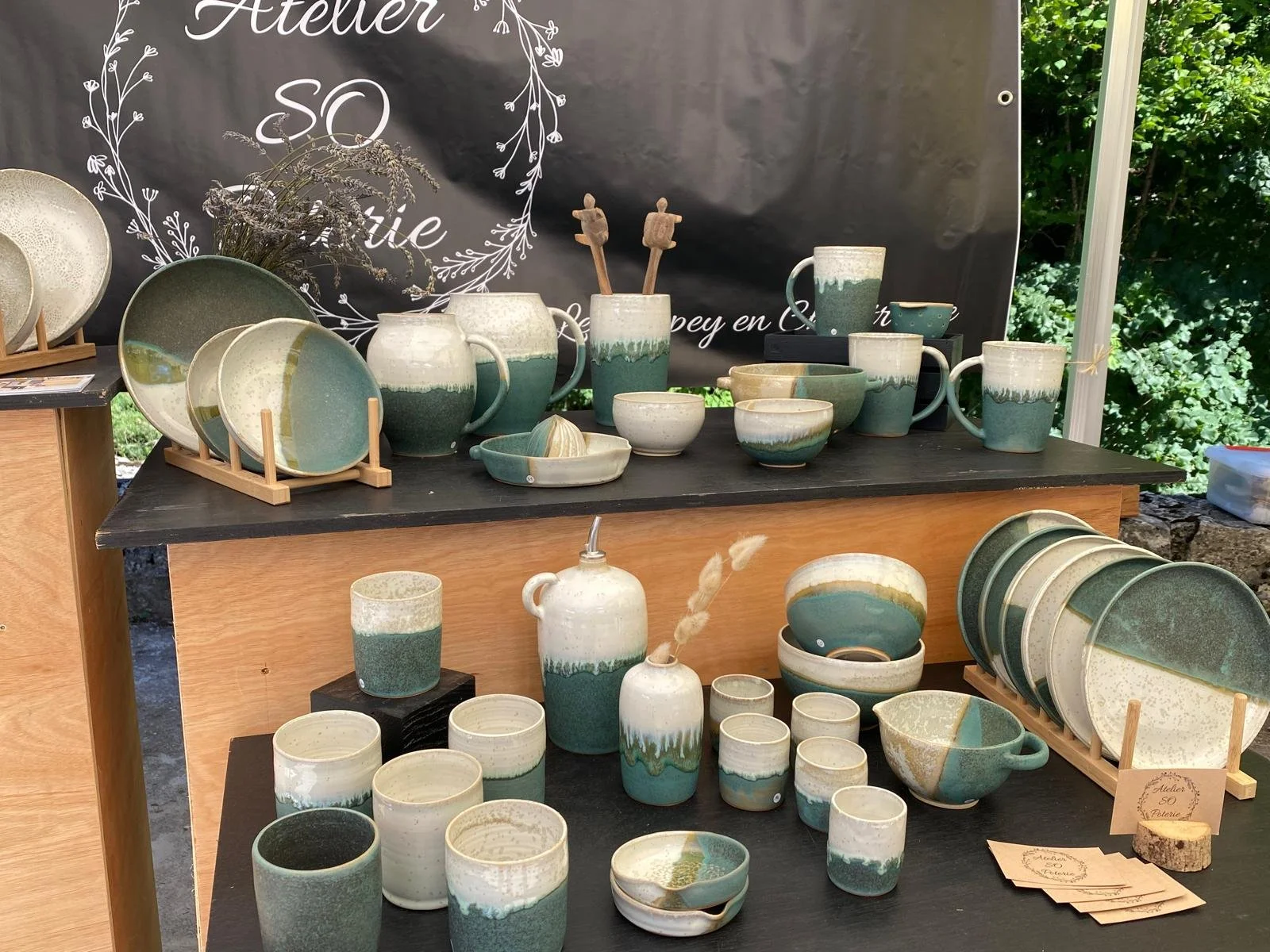 Vitrine de vaisselle en céramique verte et crème avec plats, mugs, bols, et petits contenants, décorée avec de la lavande séchée et quelques papillons en bois. Sombre fond noir avec texte "Atelier SO".