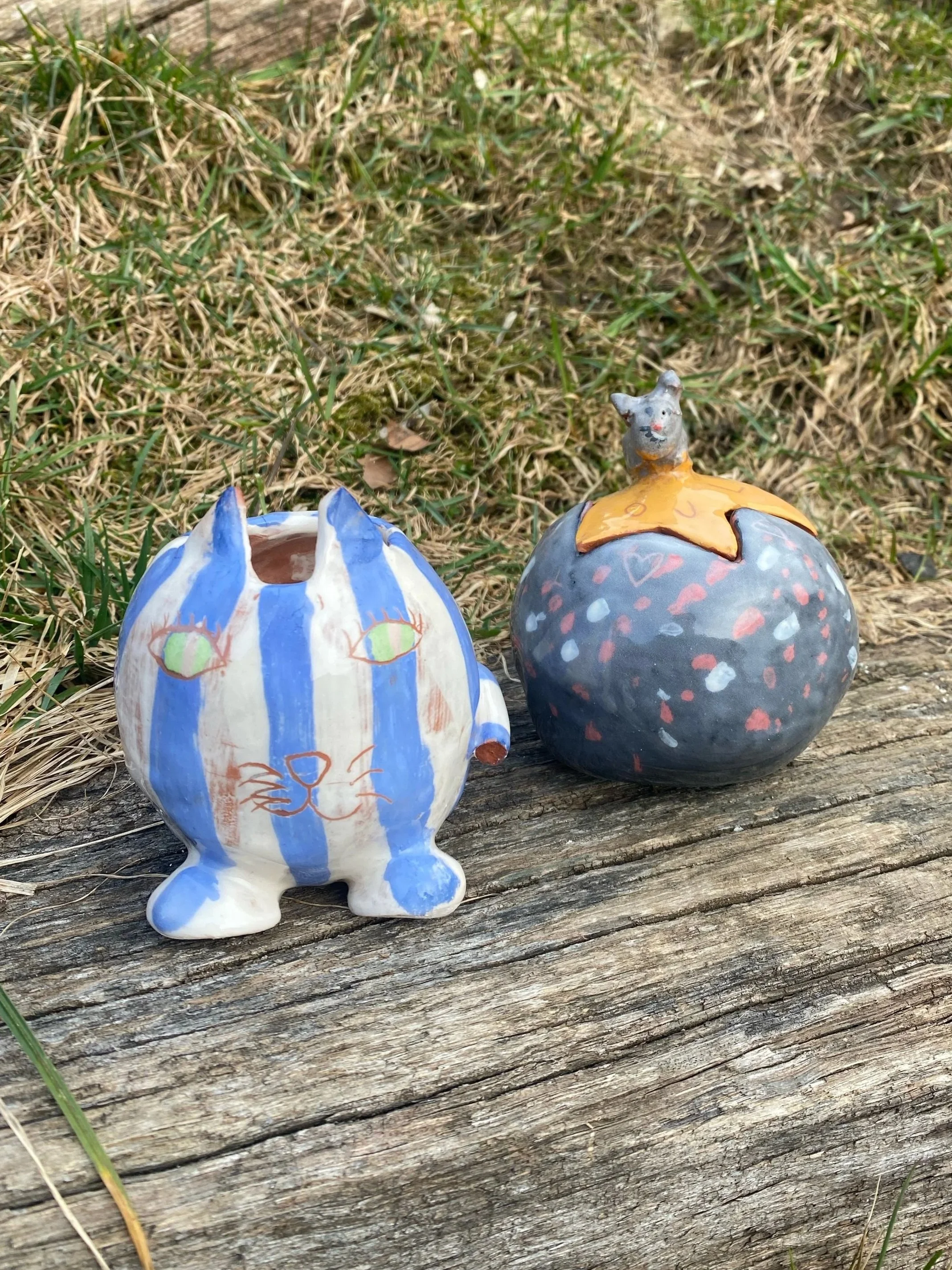 Deux figurines en céramique représentant des animaux imaginaire, placées sur un morceau de bois avec de l'herbe en arrière-plan.