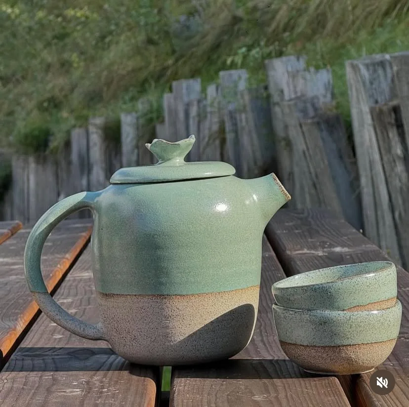 Une théière et deux tasses en grès verte sur une table en bois, en plein air.