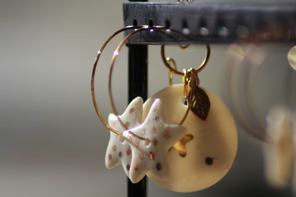 Boucles d'oreilles perchées sur un support noir, comprenant une étoile en céramique blanche avec des points dorés, une lune en céramique blanche, et une feuille en métal doré.
