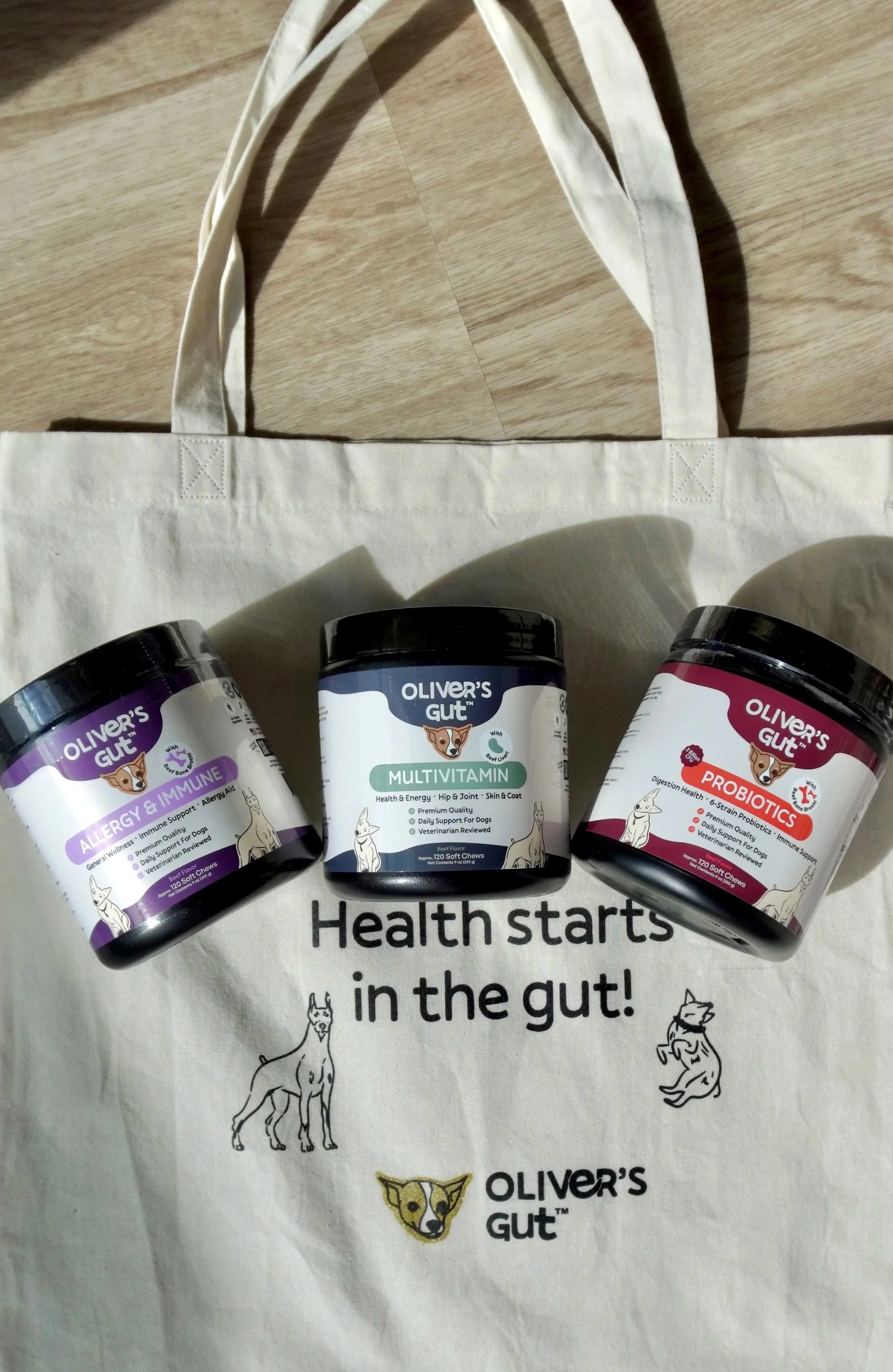 Three containers of Oliver's Gut dog supplements (Allergy & Immune, Multivitamin, Probiotics) placed on a tote bag with the slogan "Health starts in the gut!" and dog illustrations.