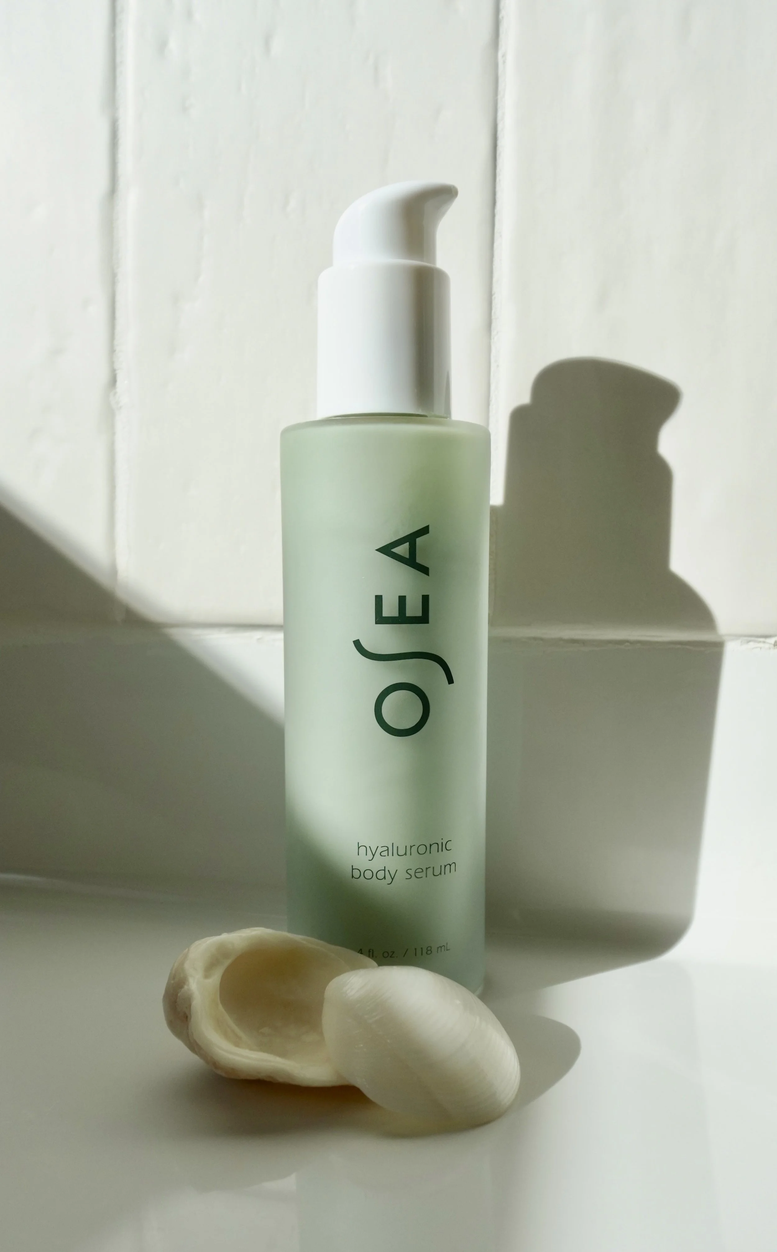 A bottle of OSEA hyaluronic body serum placed on a white surface with a seashell and a smaller white shell in front of it, casting shadows on a white tiled wall.