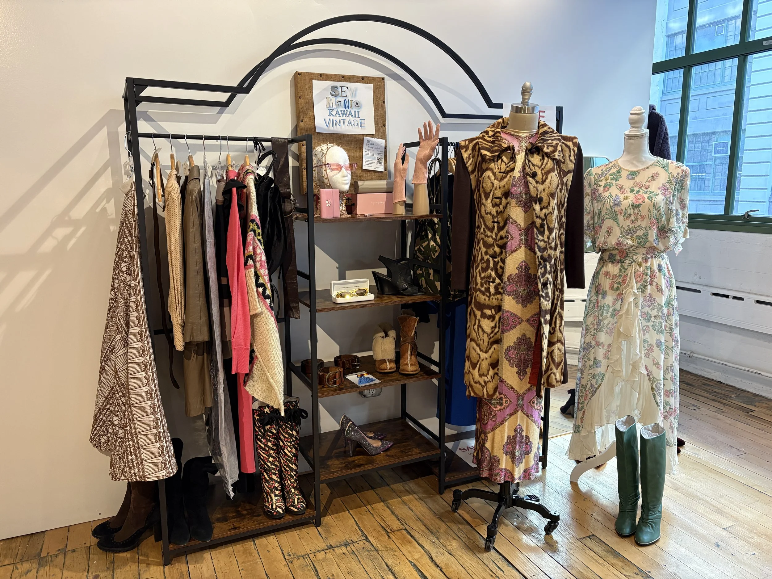 Display of vintage clothing and accessories with two mannequins dressed in floral and animal print garments, a clothing rack with jackets and dresses, shelves with shoes and jewelry, and a window showing city buildings.