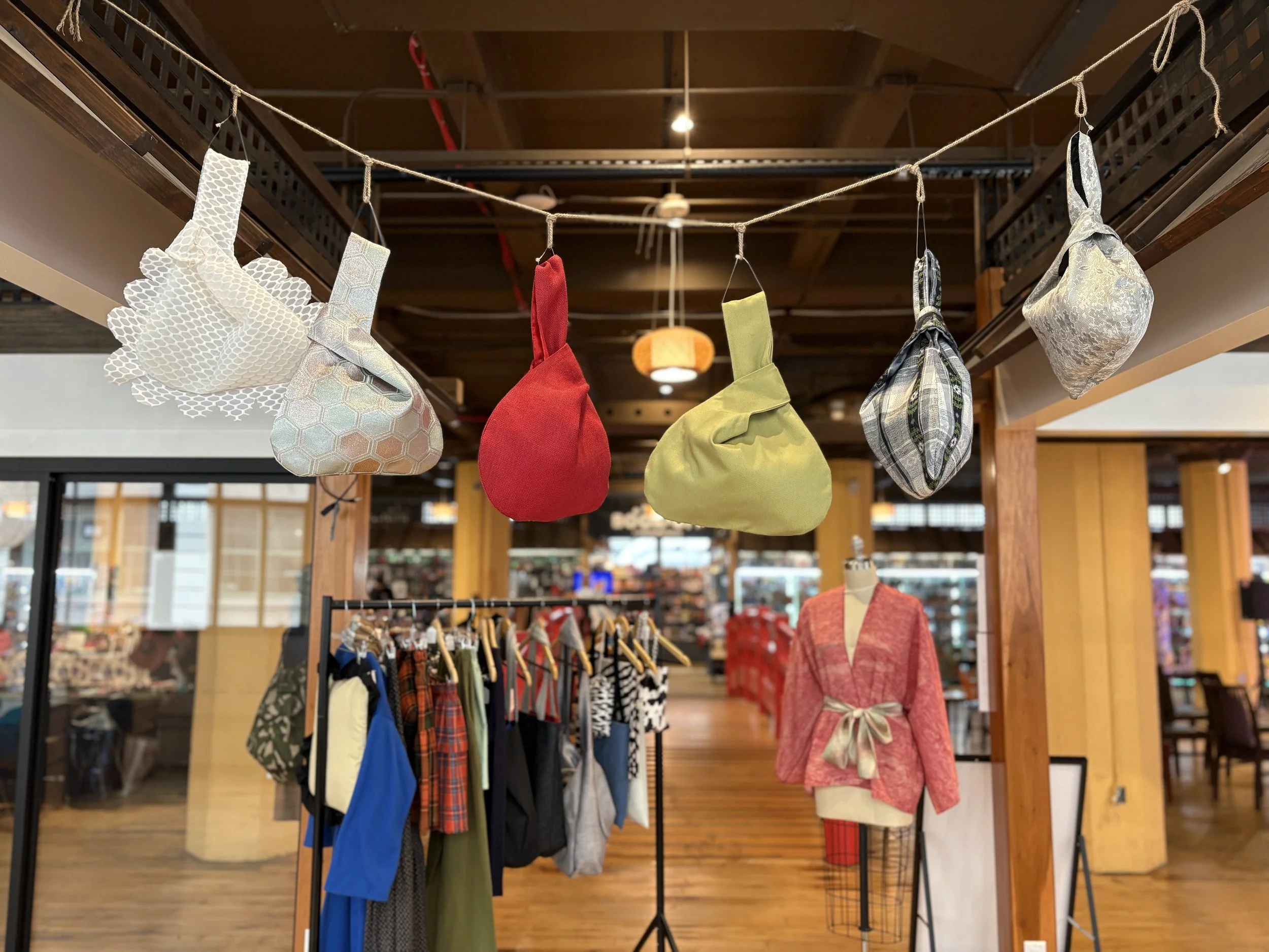 Colorful fabric pouches hanging from a string in a boutique clothing store with racks of clothes and a mannequin in the background.