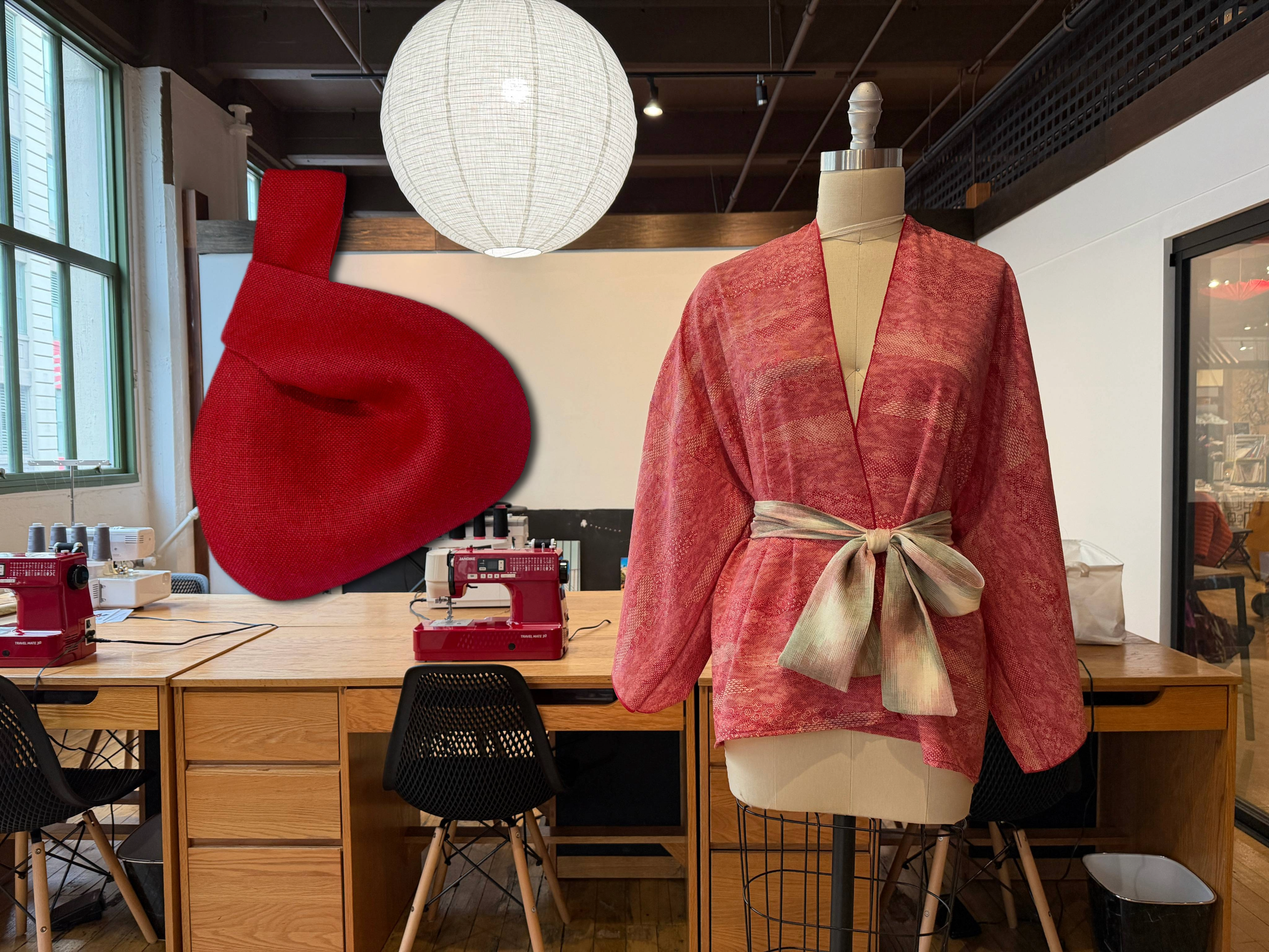A fashion display featuring a red fabric sculpture resembling a mitten on the left and a mannequin on the right dressed in a pinkish-red kimono-style jacket with a waist tie, inside a retail or craft store.