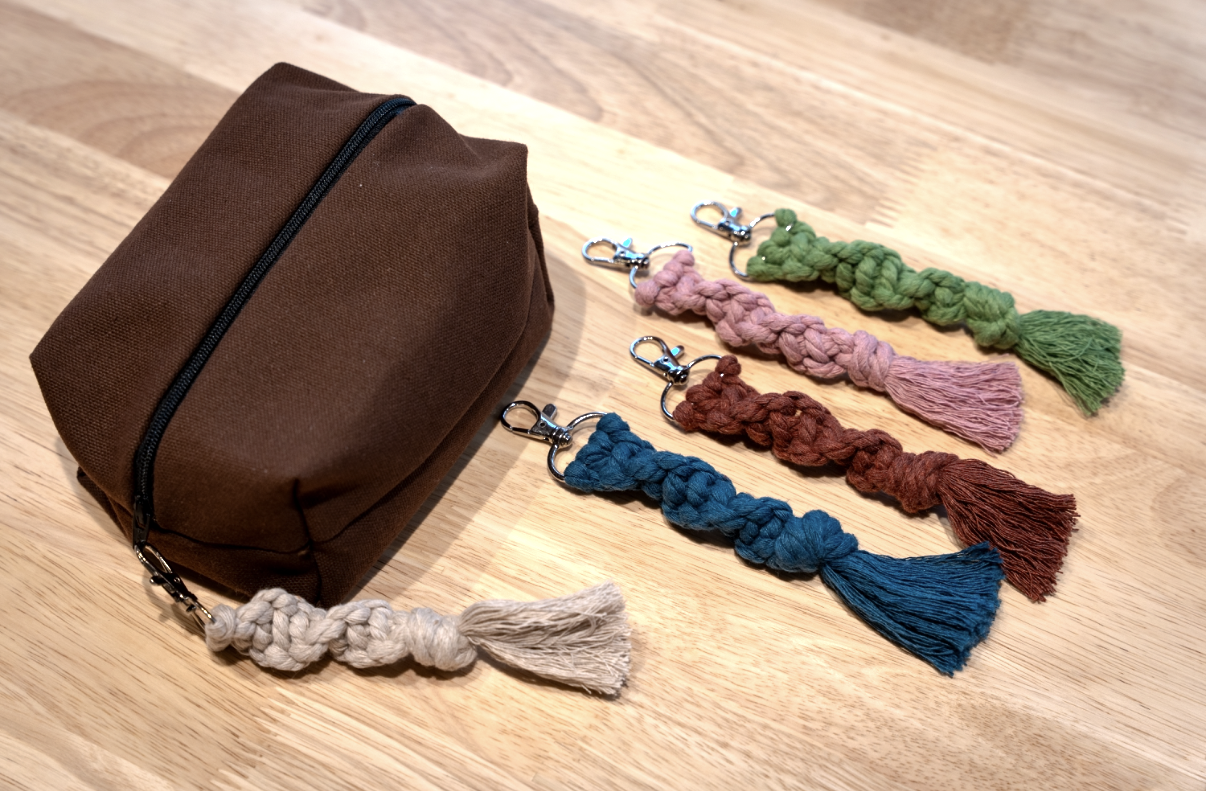 Seven colorful braided tassel charms laid out next to a brown fabric zippered bag on a wooden surface.
