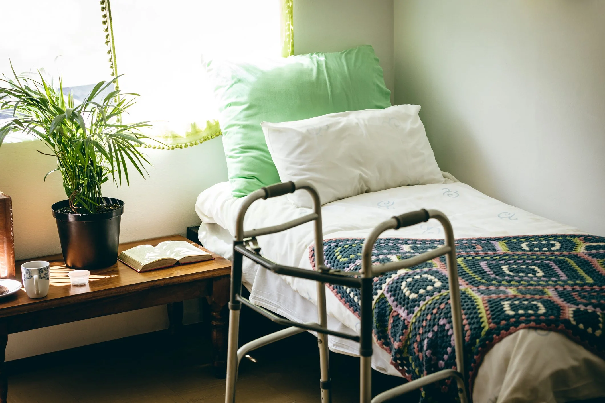 A bedroom with a bed, green and white pillows, a walker next to the bed, a wooden nightstand with a potted plant, a book, a cup, and a small container, and a window with green curtains.