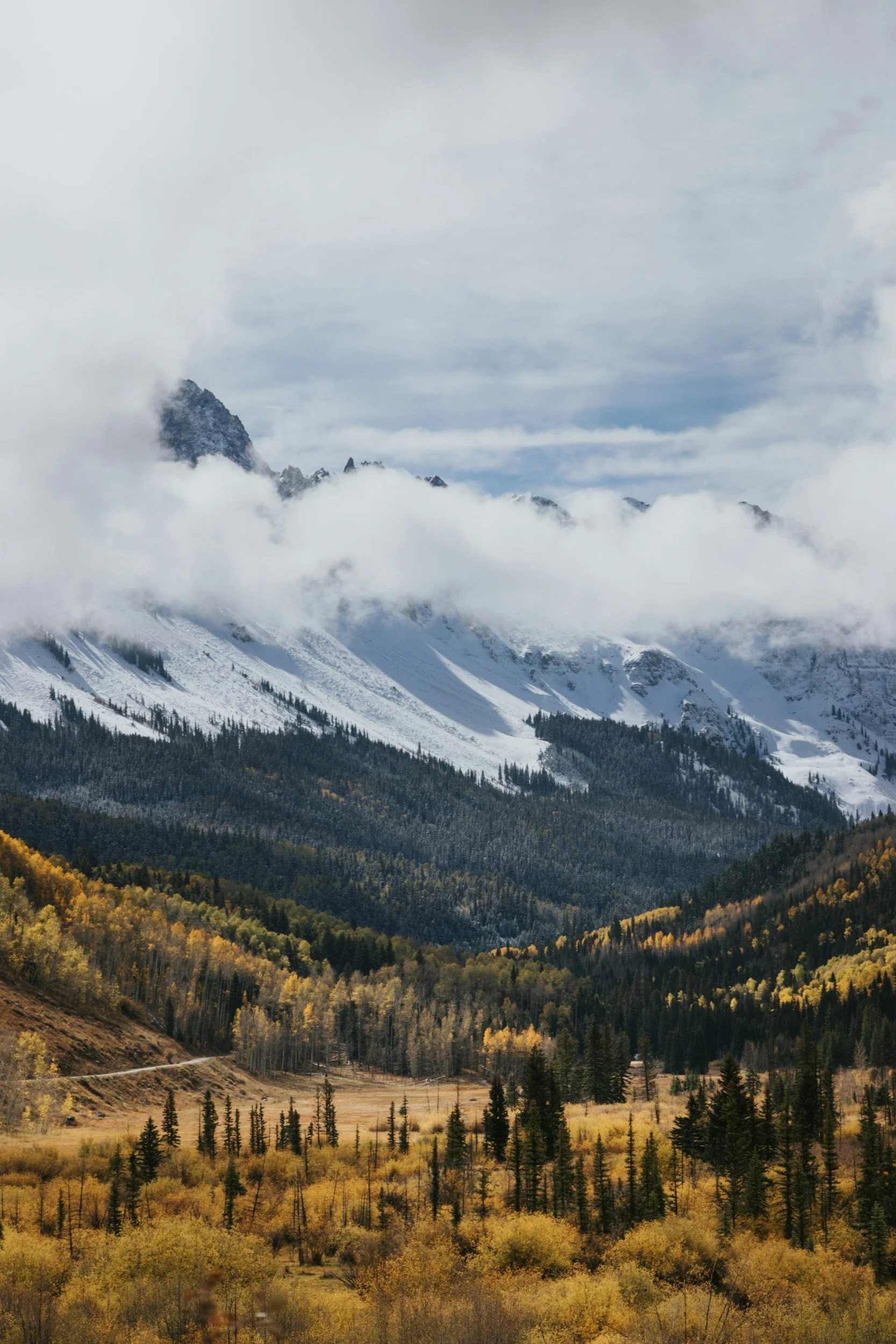 Scenic mountain landscape with snow-capped peaks, partially covered by clouds, surrounded by forested slopes and golden autumn foliage.