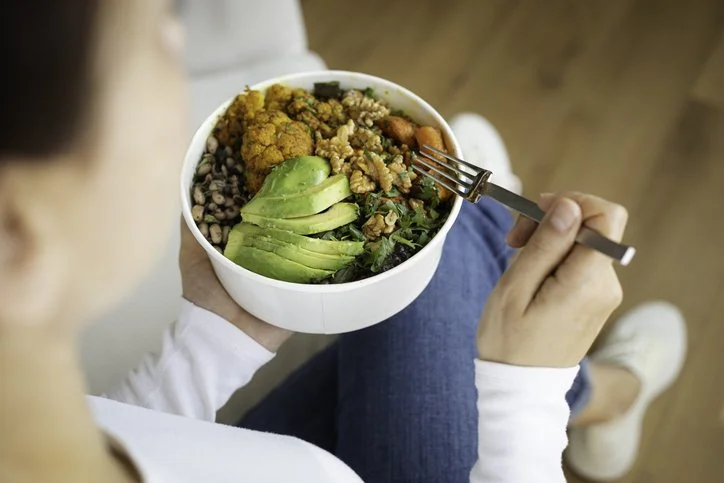 Person sitting with legs crossed, holding a bowl of food in their lap, and using a fork to pick up a piece of tomato.