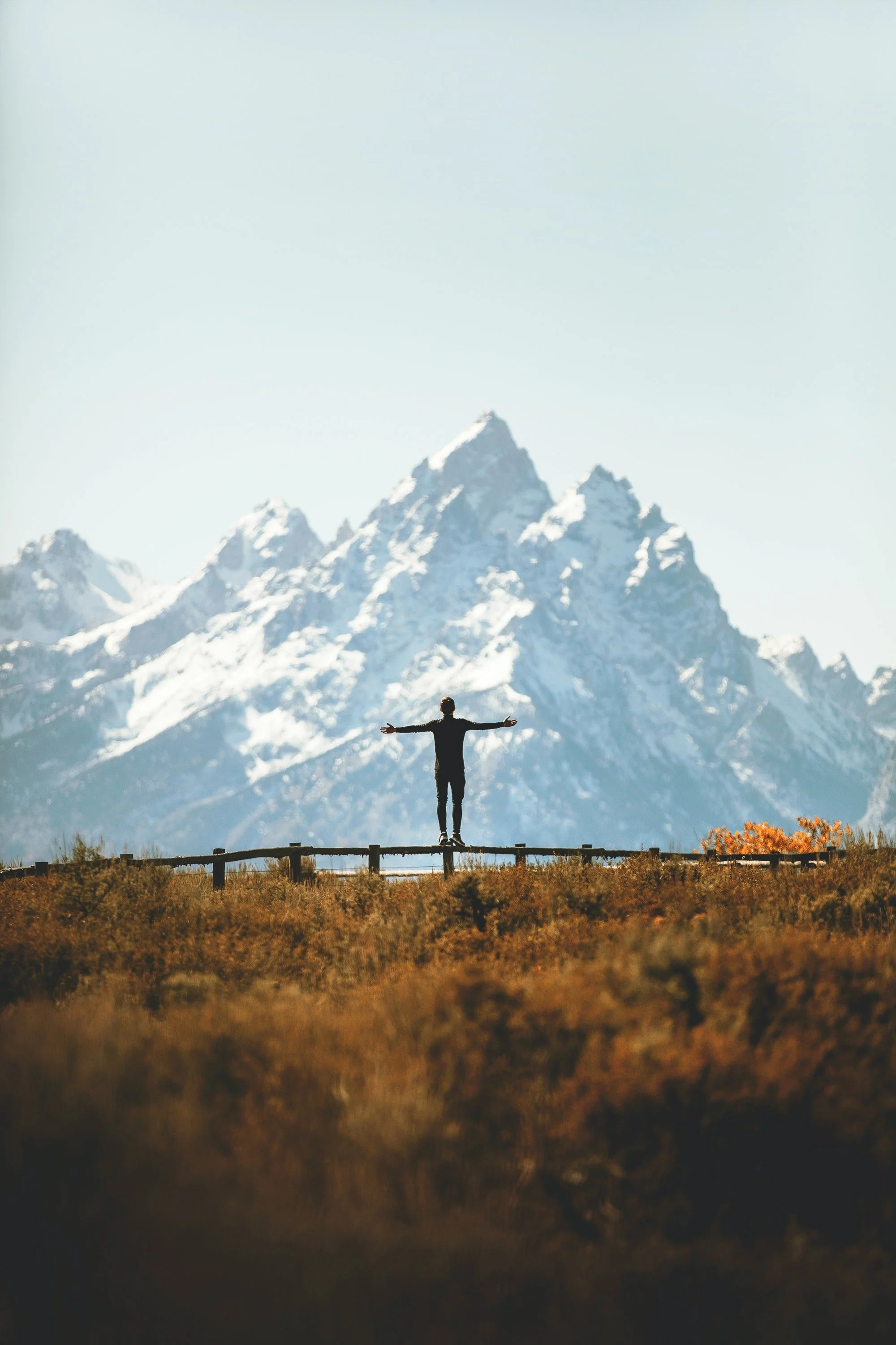 A person standing on a wooden railing with arms outstretched, facing snow-capped mountains in the distance.