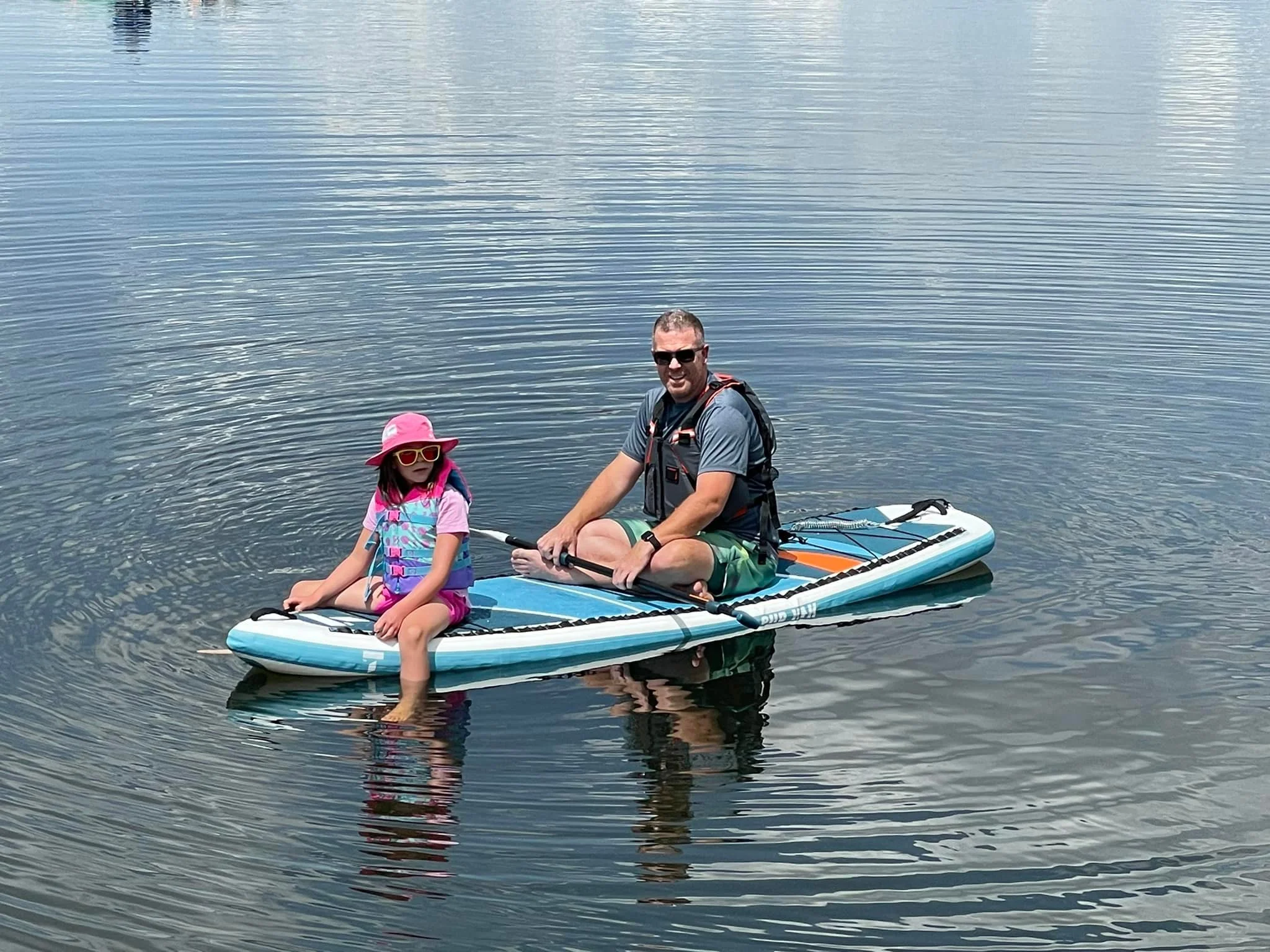A man and a young girl sit on a paddleboard in calm water. The girl wears a pink hat, pink sunglasses, and a life vest, while the man wears sunglasses, a gray shirt, and a life vest, and is holding a paddle.