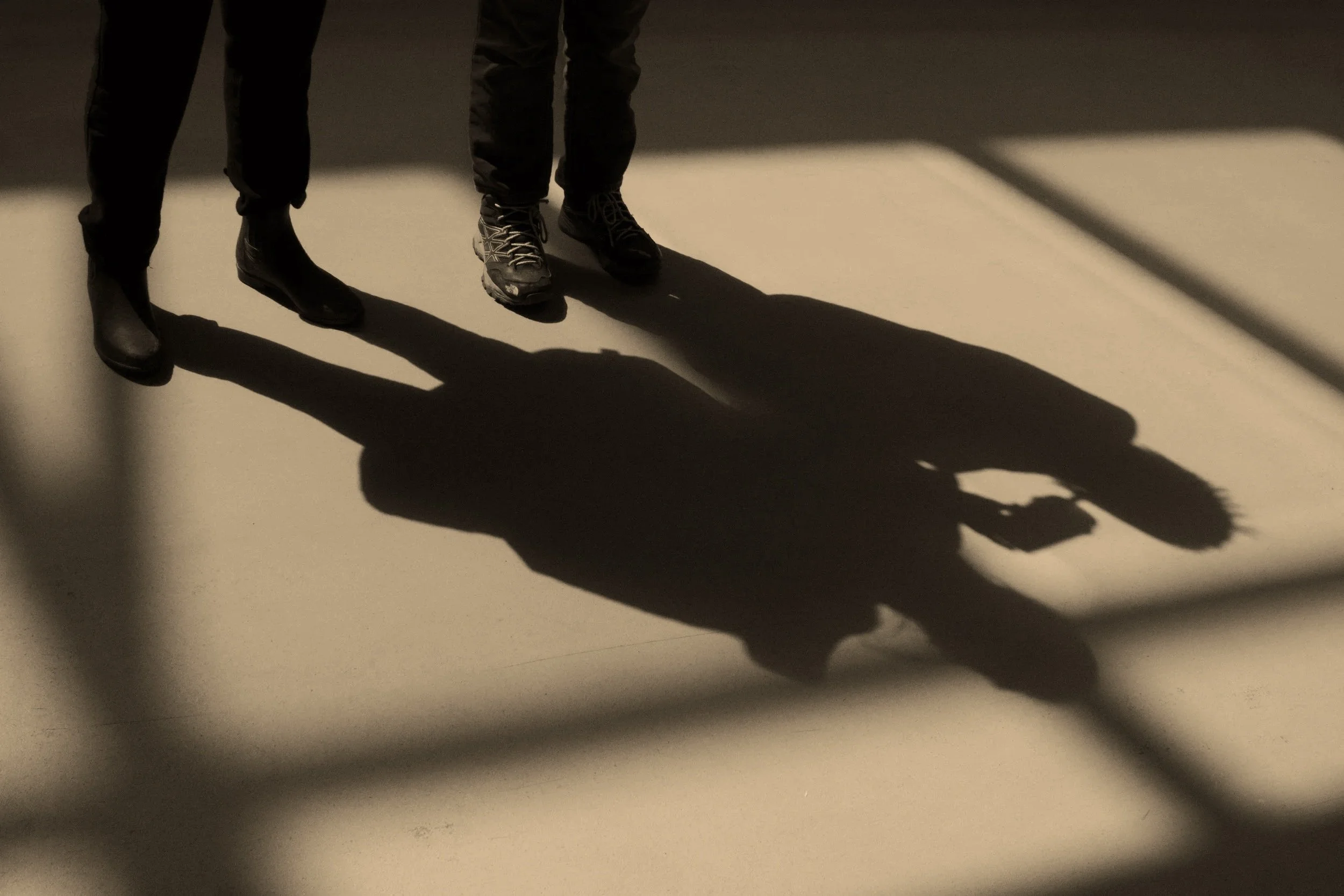 Shadow of two people standing on a beige floor, with partial view of their legs and shoes, and shadows cast by bright light coming from the side.