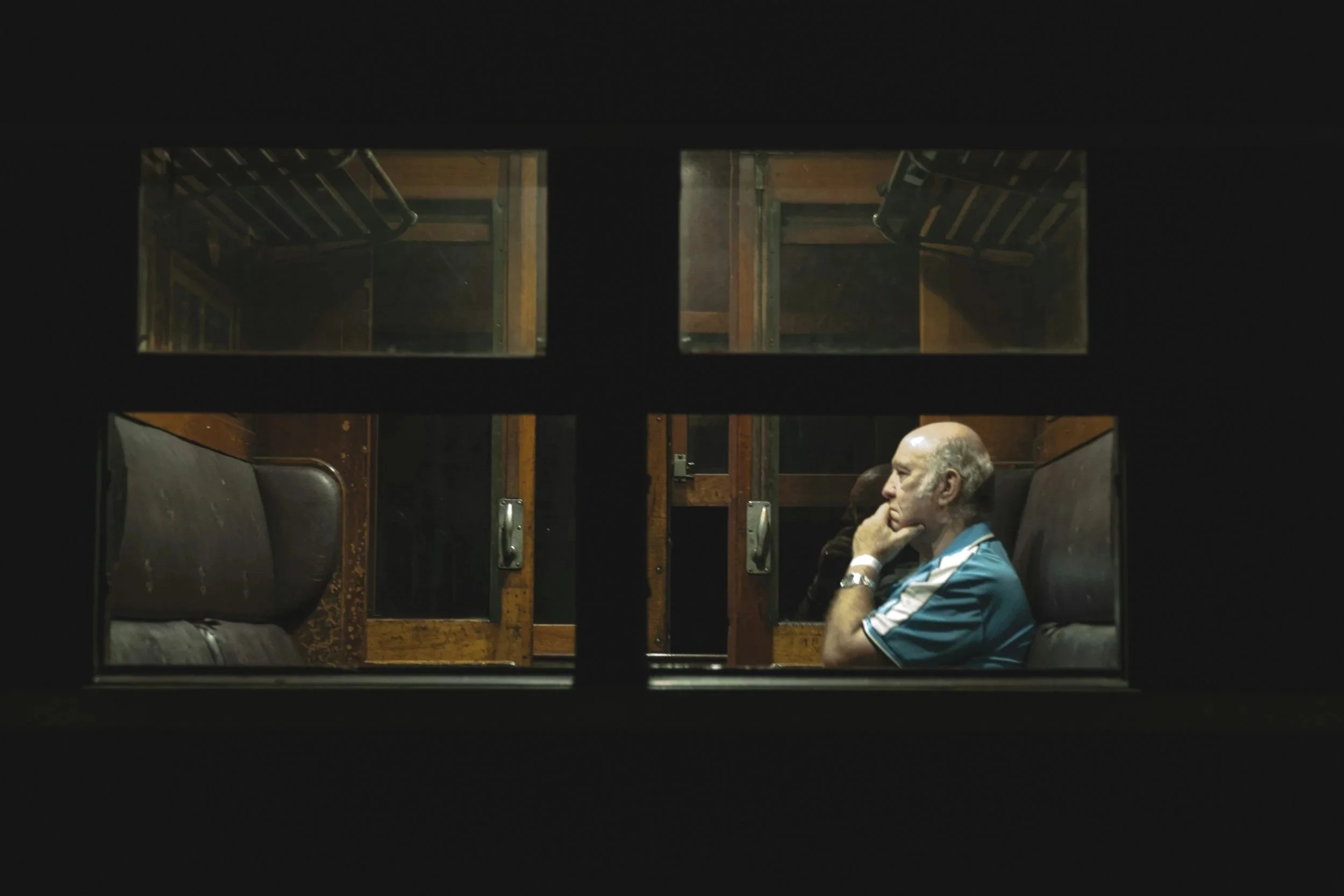 An elderly man with a bald head and a mustache, wearing a blue sports shirt, sitting on a bench inside a train, seen through a window.