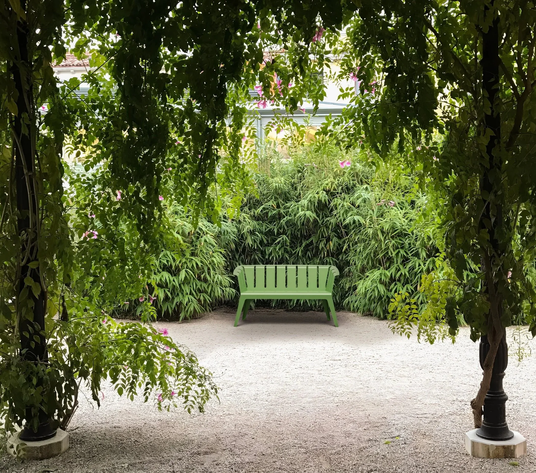 Grüner Gartenbank inmitten von üppigem Grün, umgeben von Büschen und Bäumen, in einem Garten mit Sandboden, Blick durch einen Torbogen aus Pflanzen.