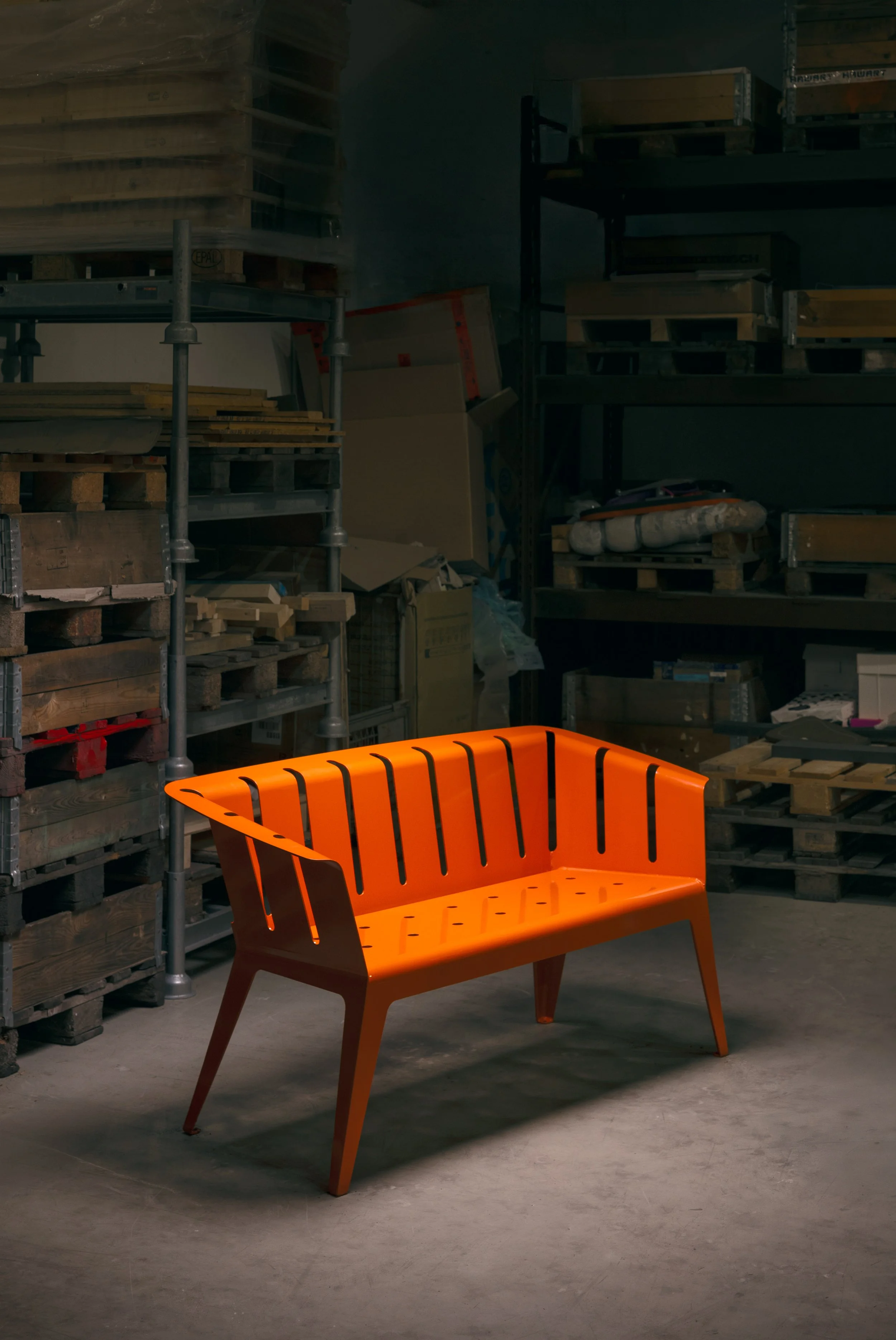 Ein orangefarbener Gartenbank steht in einem Lagerraum mit Regalen voller Baumaterialien und Verpackungen.