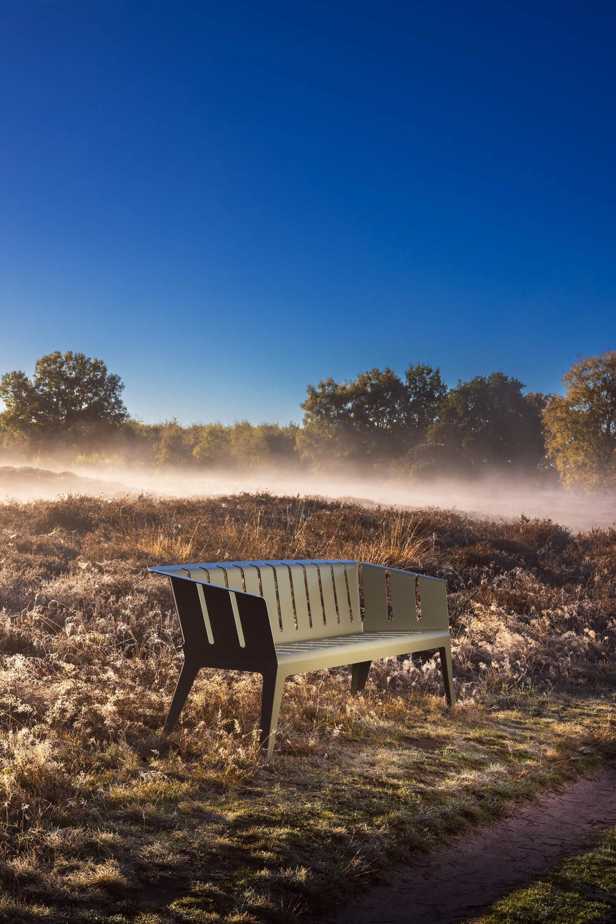 Eine einzelne Bank auf einer Feldwiese bei Morgendunst, im Hintergrund Bäume und ein klarer blauer Himmel.