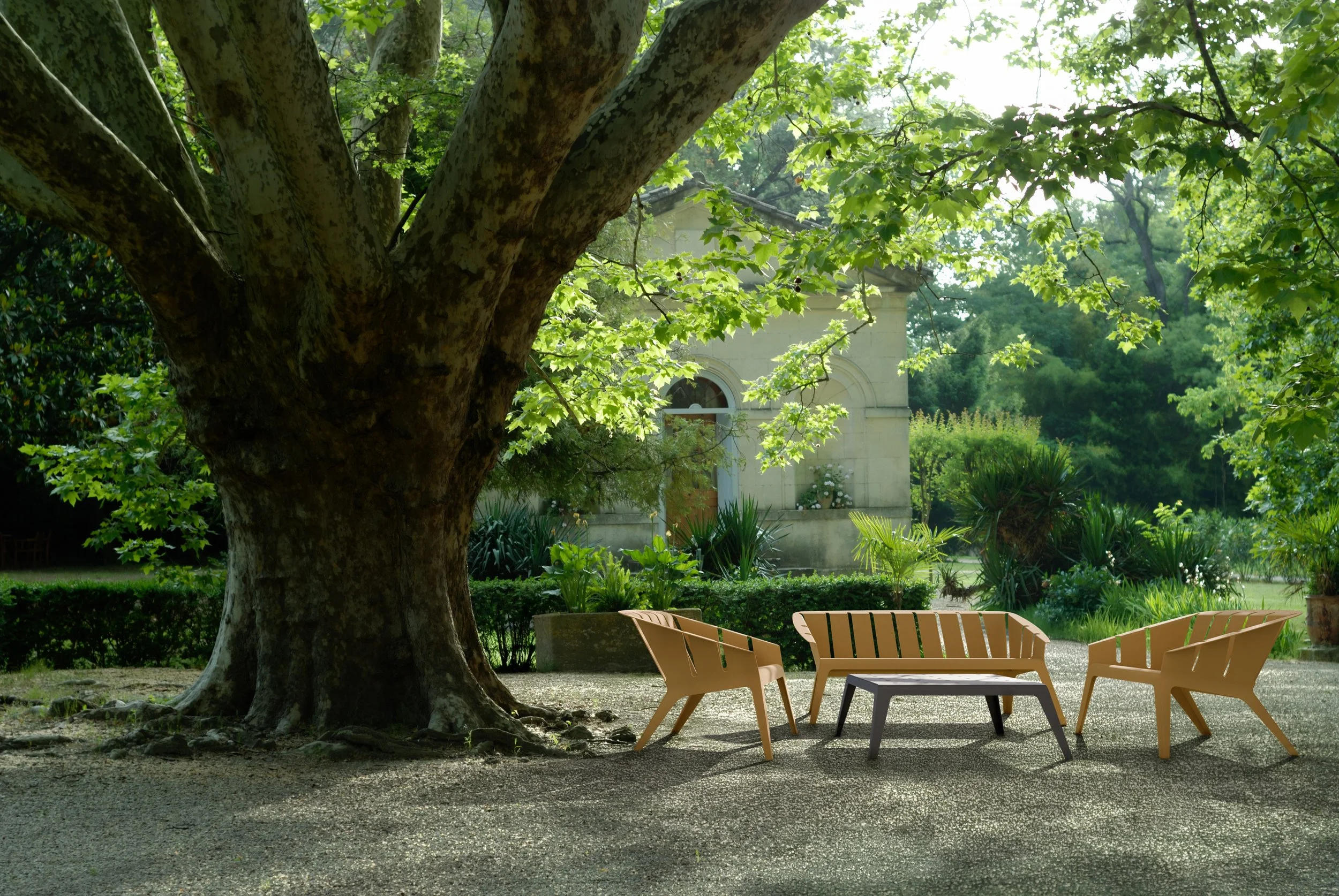 Ein großer Baum mit breitem Stamm und grünen Blättern, im Hintergrund eine kleine weiße Kapelle, umgeben von Rasen und Büsche, mit Gartenmöbeln aus Holz auf einem Kiesweg im Vordergrund.