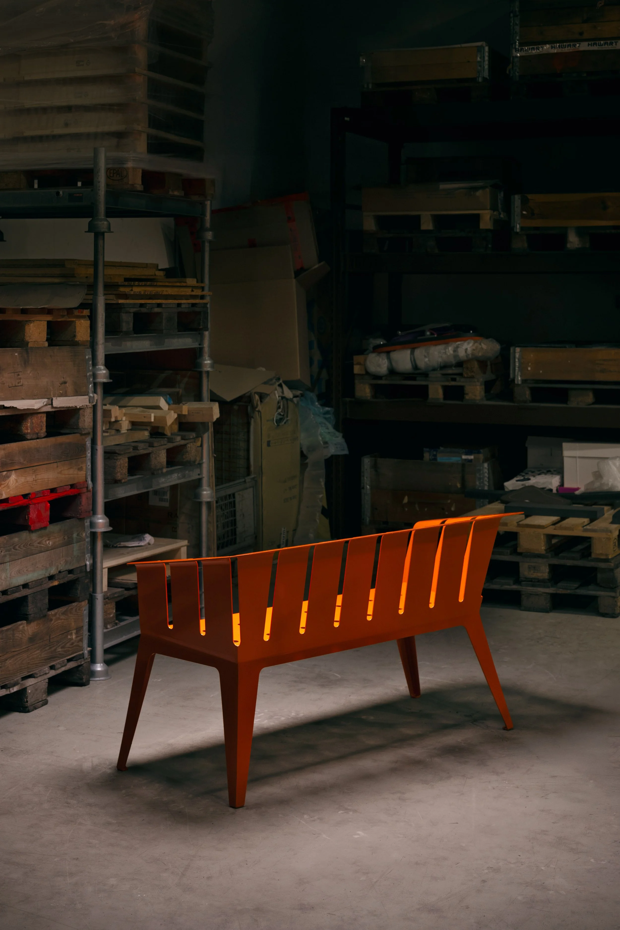 Ein orangefarbener Holzbank in einem Lagerraum mit Werkzeug, Holz und Paletten im Hintergrund.