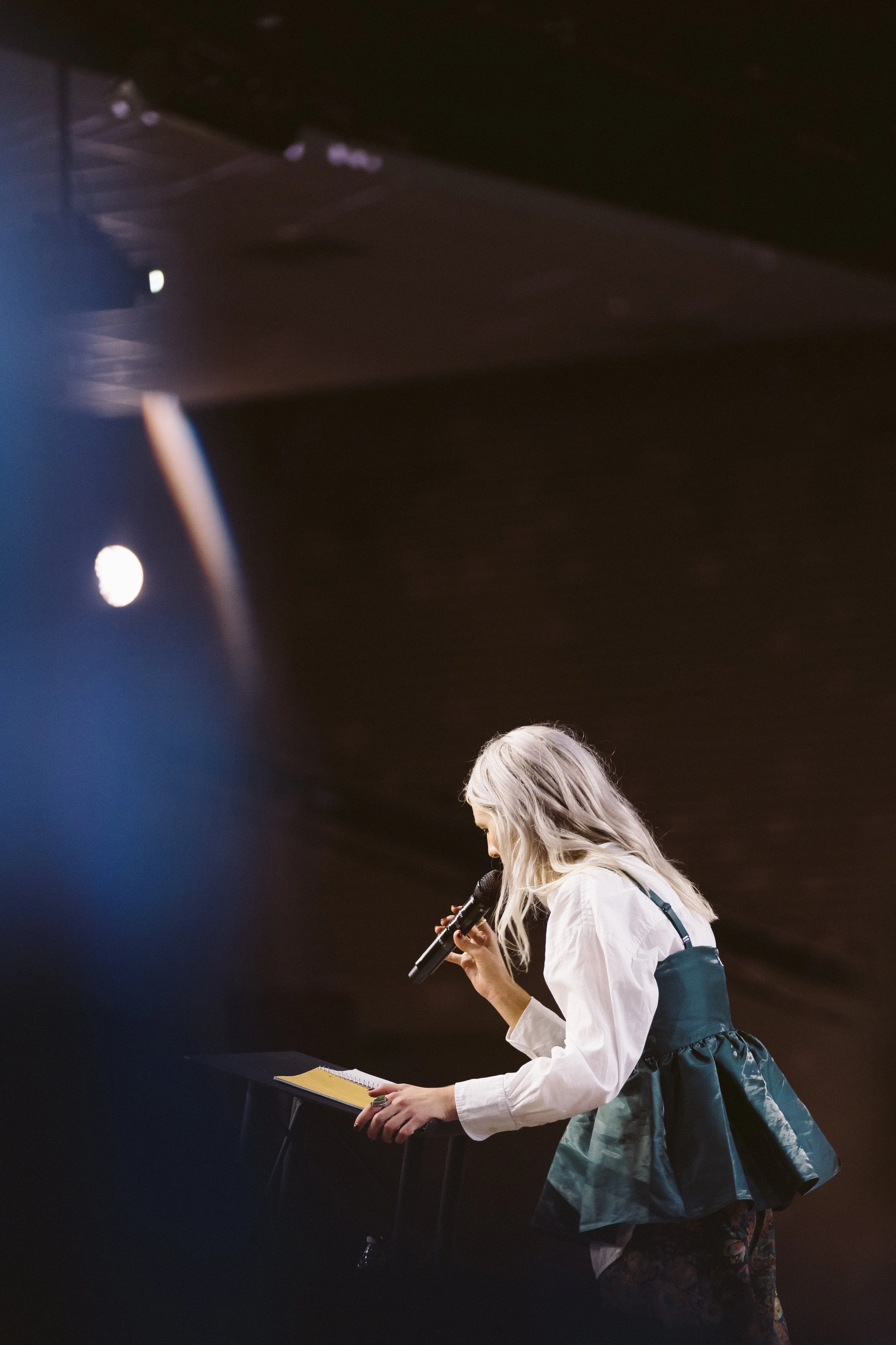 A woman with long, blonde hair standing on stage, holding a microphone and reading from a notebook.