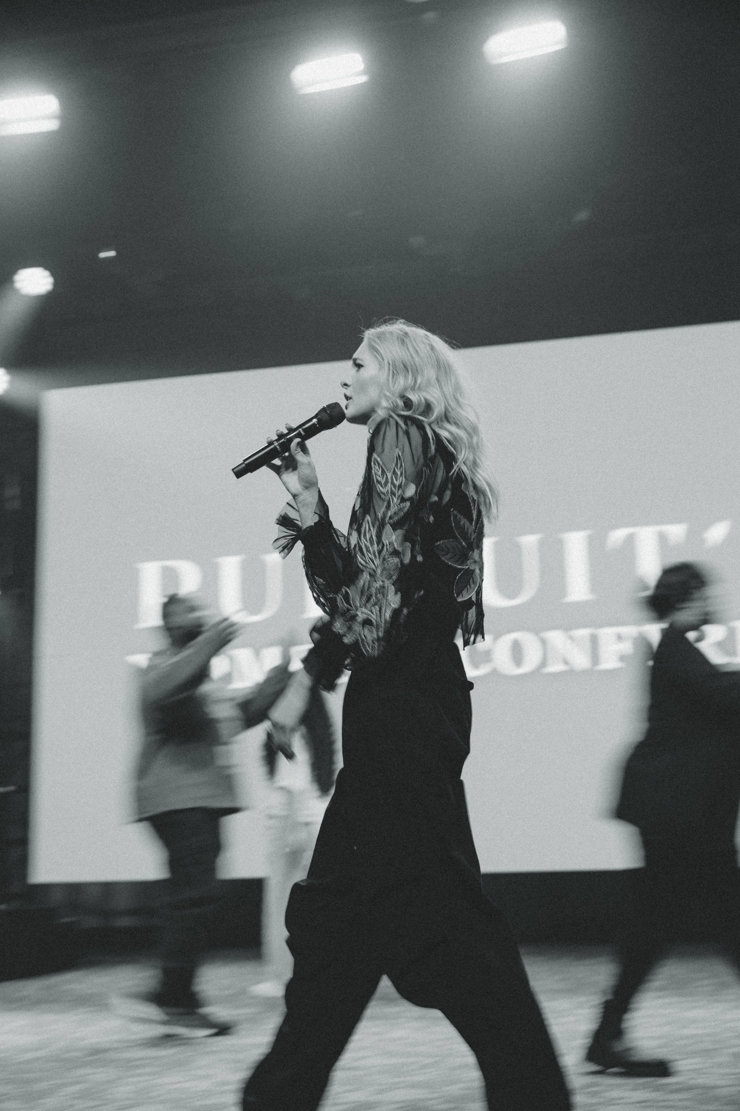 A woman with wavy blonde hair singing into a microphone on stage during an event, with blurred people in the background and a large screen displaying text.