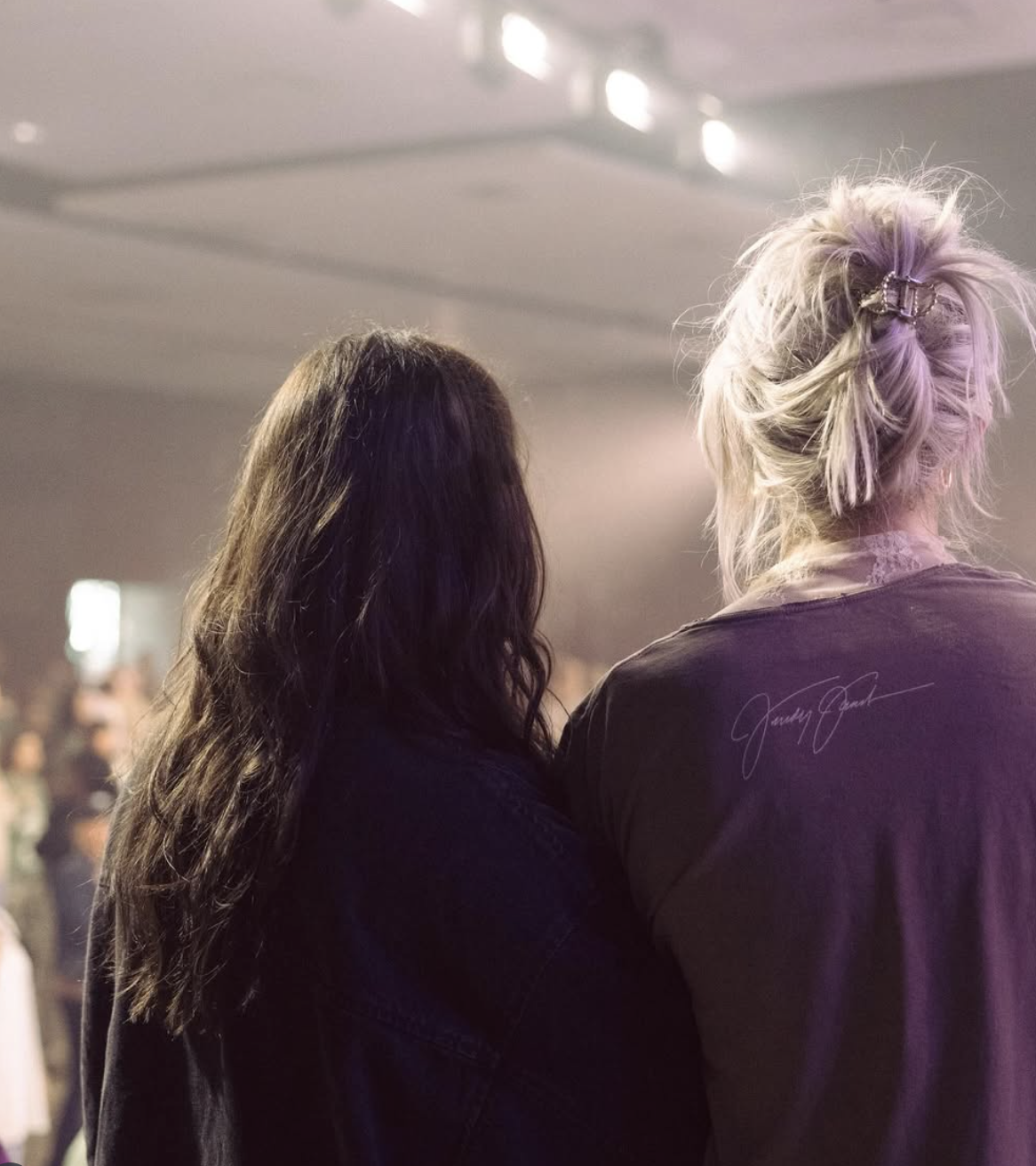 Two women with long hair, one with dark hair and the other with platinum blonde hair tied up, are standing close together at an indoor event with many people in the background.