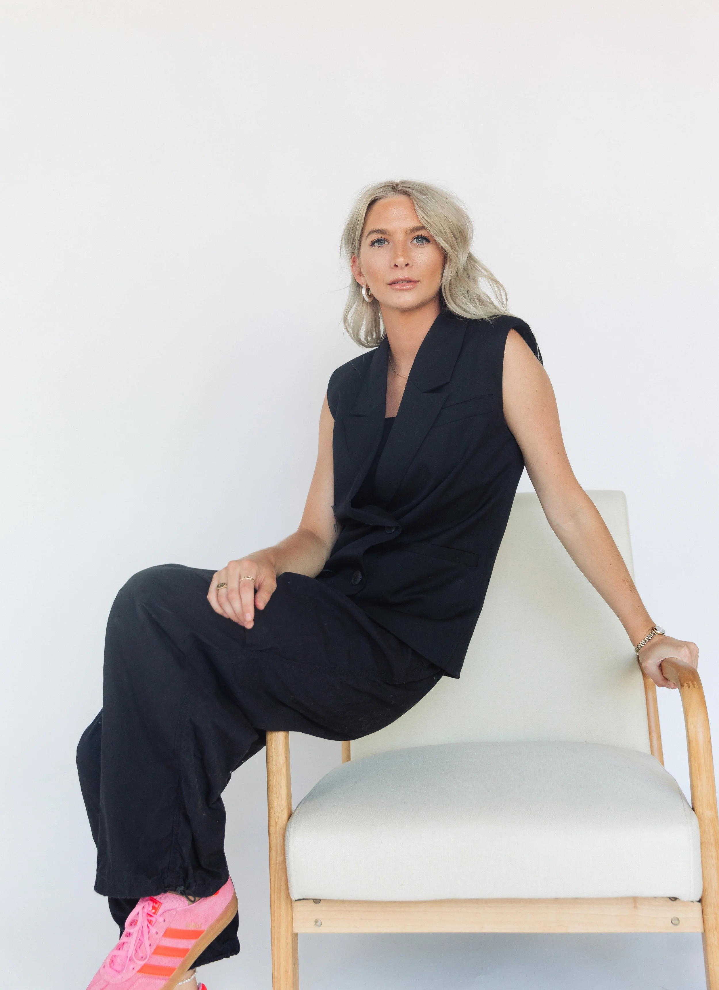 A woman with blonde hair, wearing a sleeveless black blazer, black pants, and pink sneakers, sitting on a wooden chair with her right hand resting on the chair's armrest.