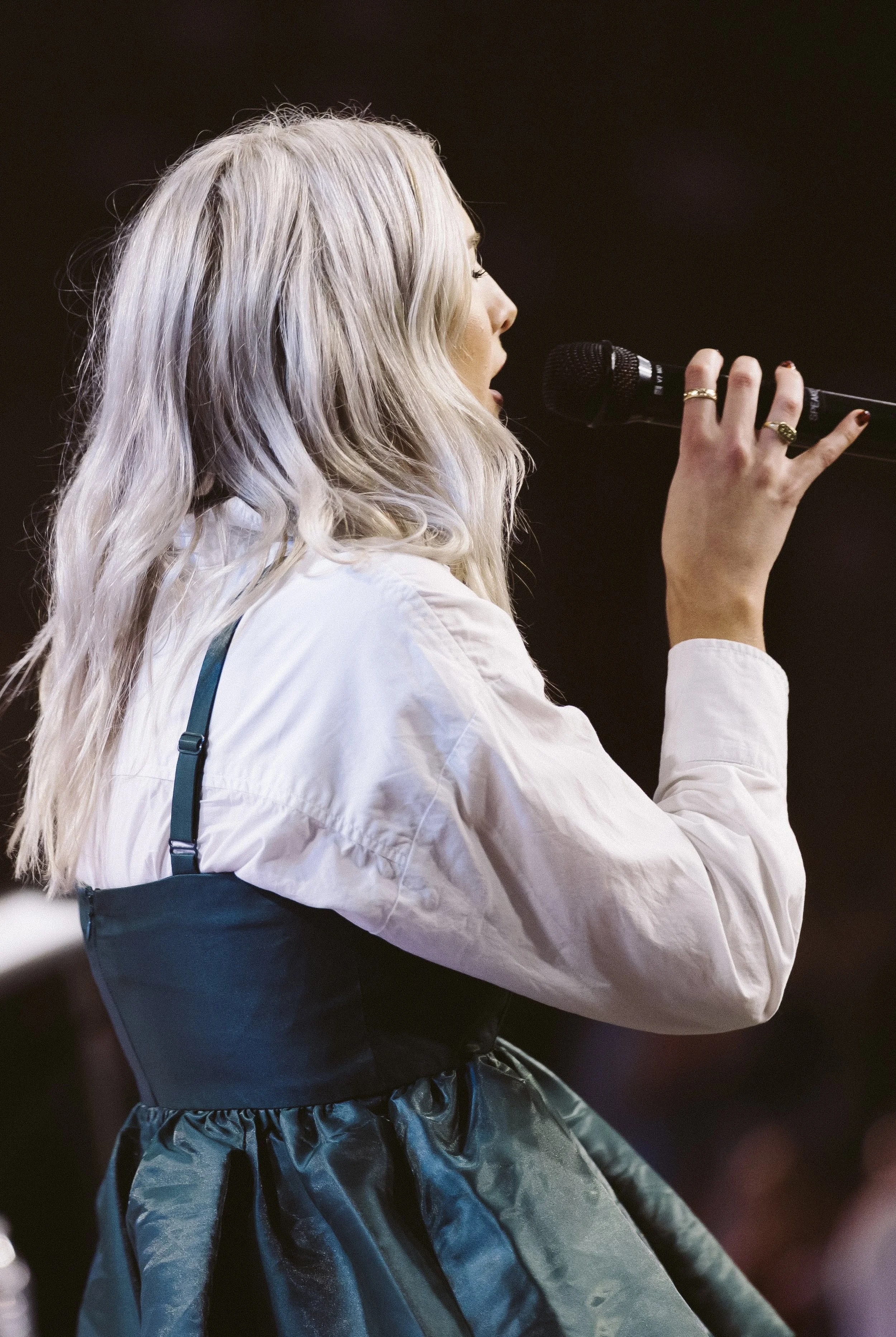 A woman with long blonde wavy hair singing into a microphone on stage.
